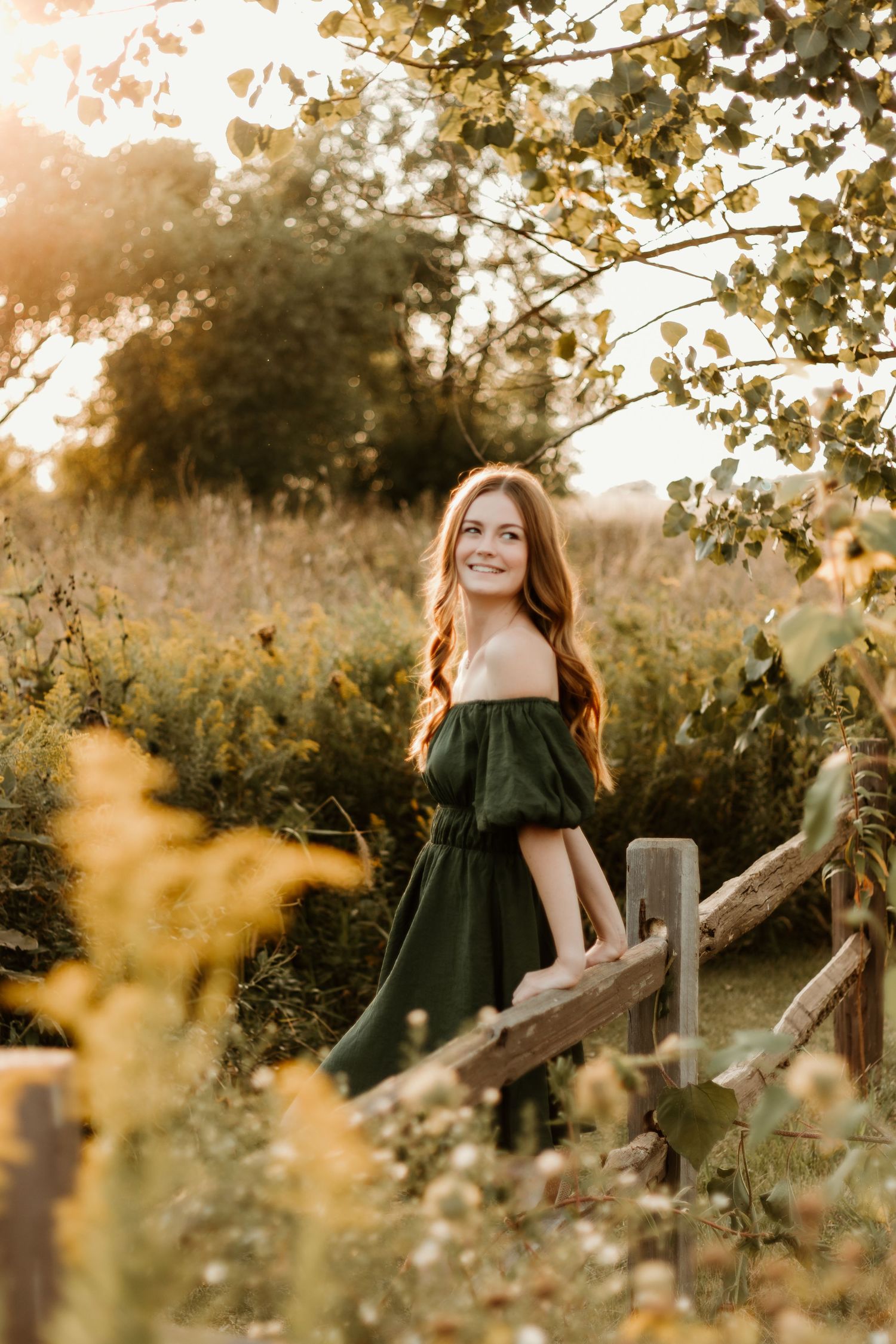 High school senior poses against a wooden fence in front of wildflowers during a senior photo session captured by central il senior photo