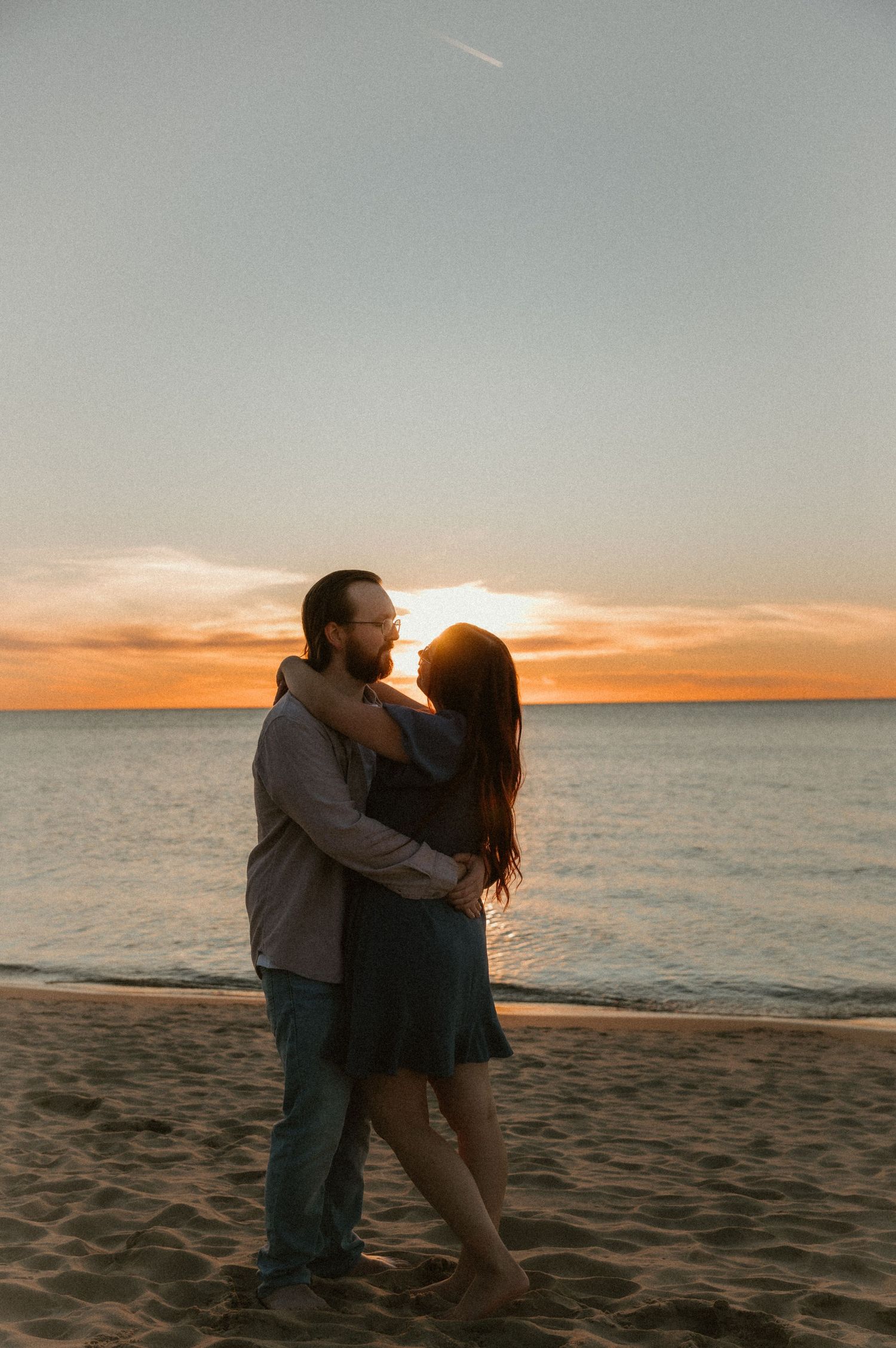 Romantic sunset beach cuddle captured in golden light during an engagement session. Couple wraps arms around each other showing their love