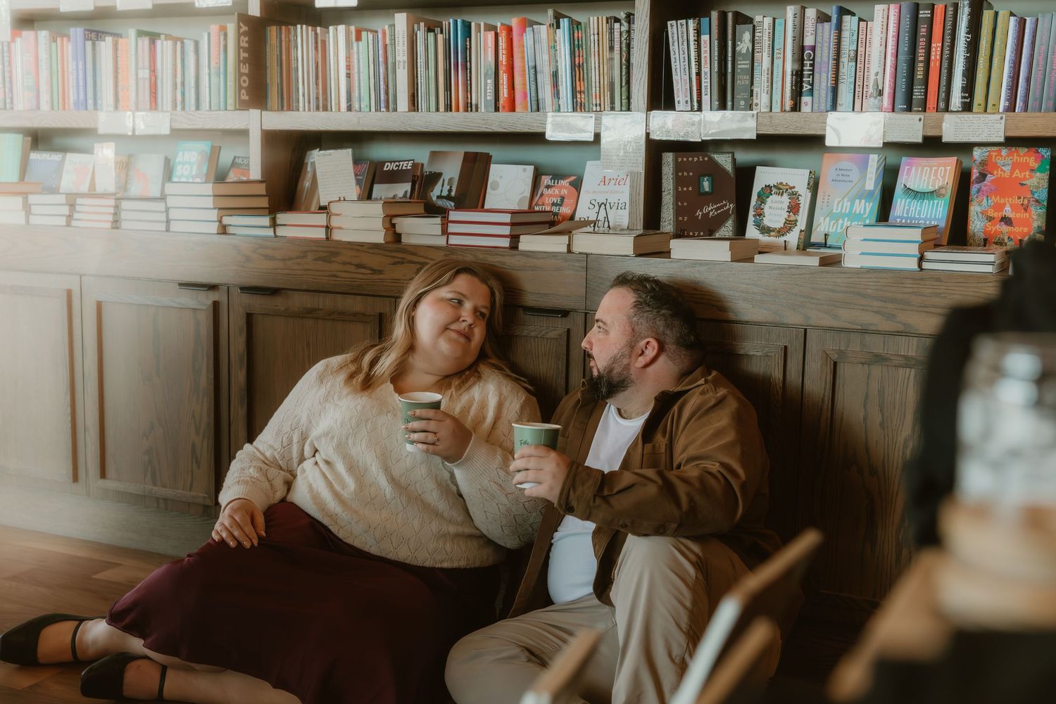 Couple sits in a bookstore sharing a cup of coffee with eachother and chatting about their wedding while photographer captures their photos