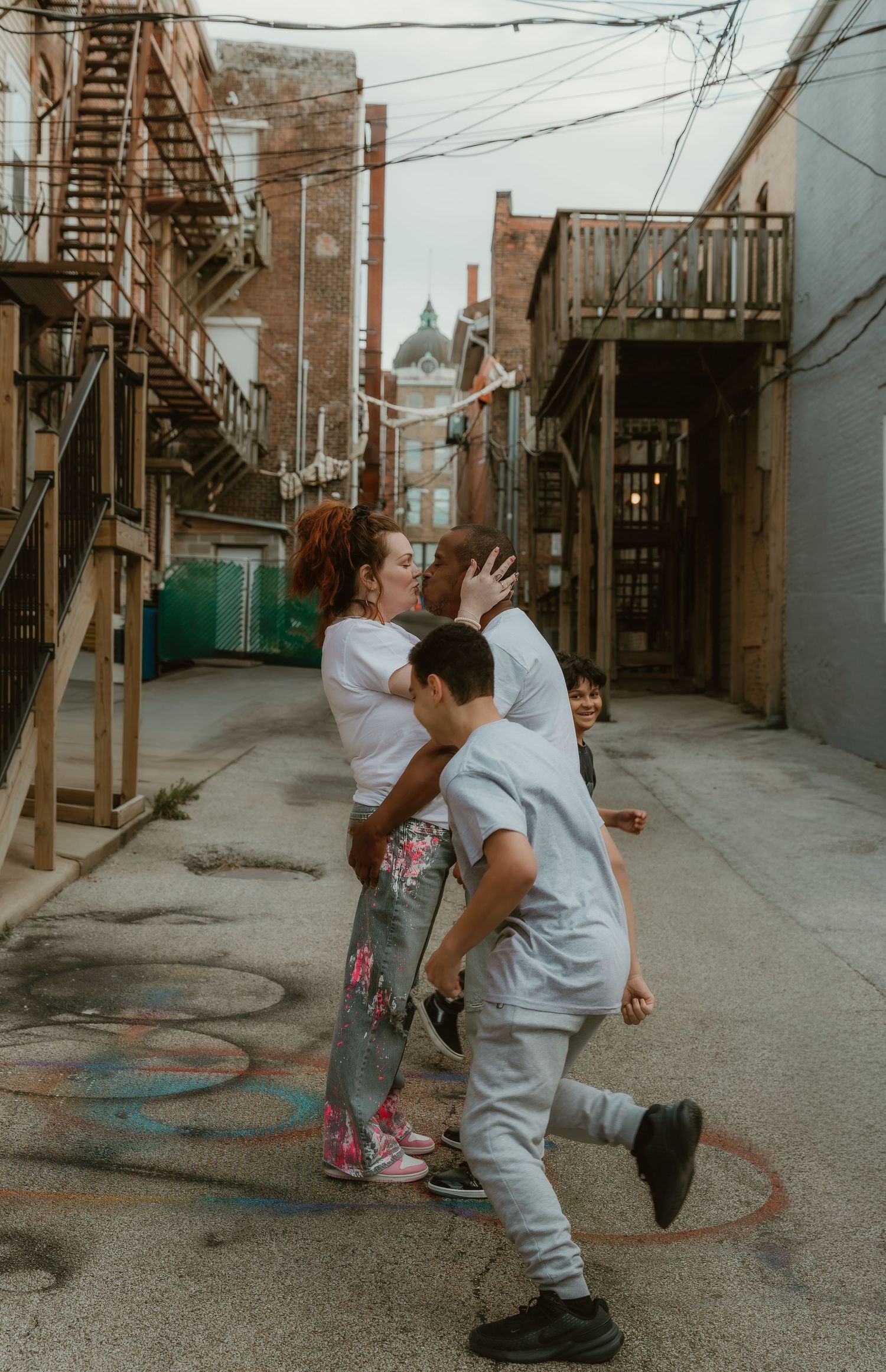 Couple exchanges a smooch while their kids run around them in Downtown Bloomington, IL during a family session