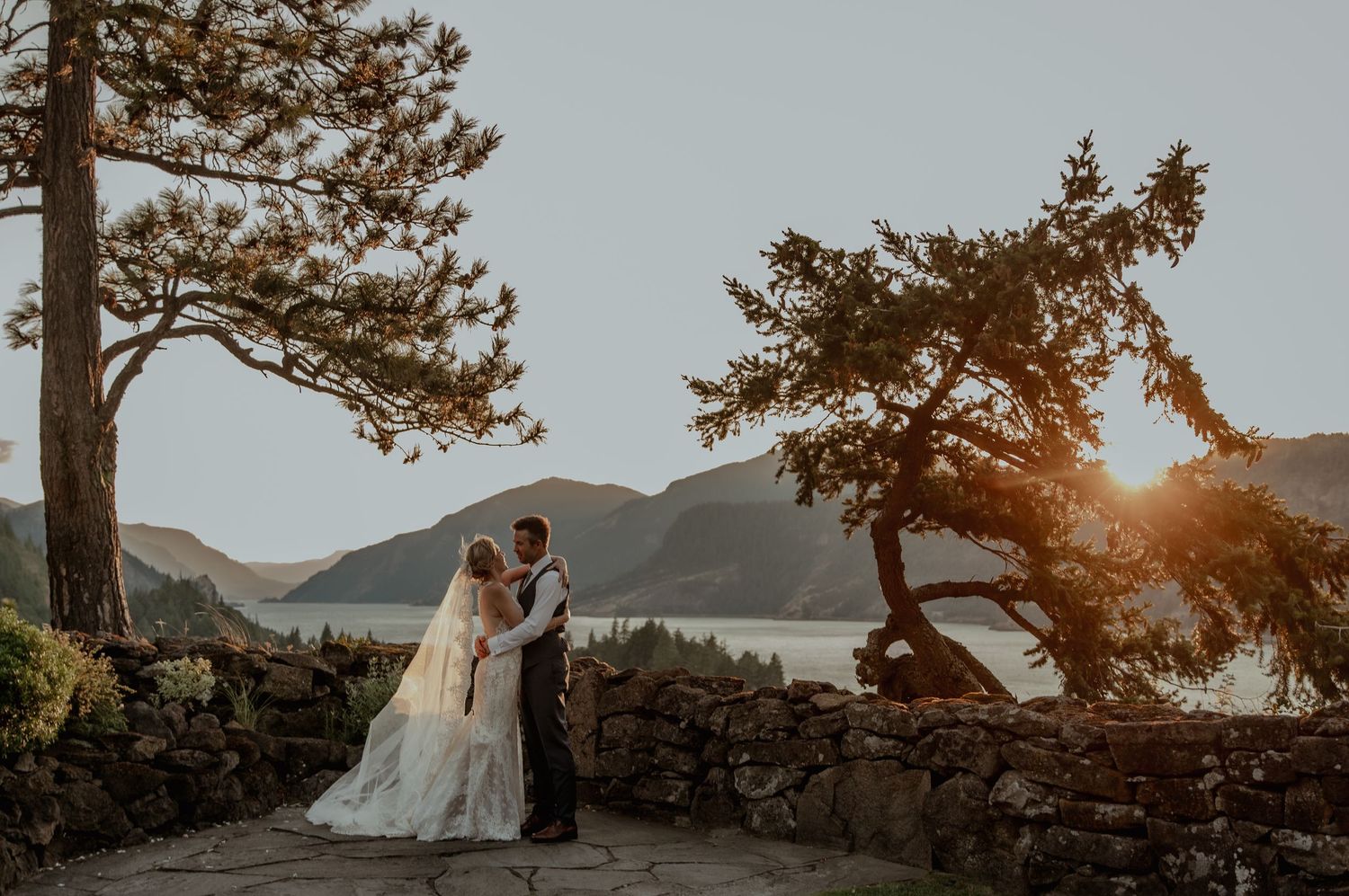 Bride and groom share a romantic moment at sunset by a stone wall with mountain lake views.