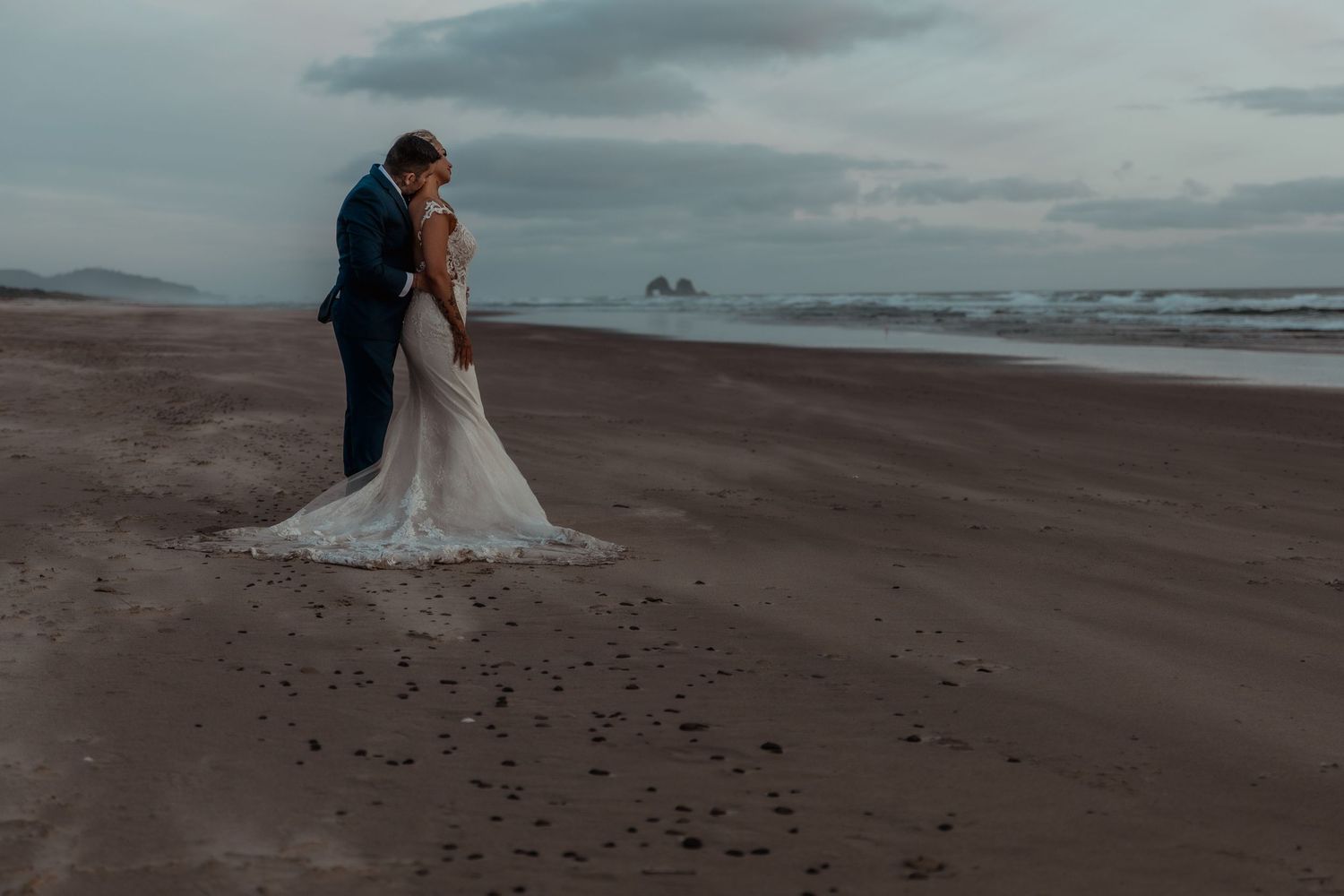 A couple embraces on a moody beach with dark clouds overhead and ocean waves breaking in the background.