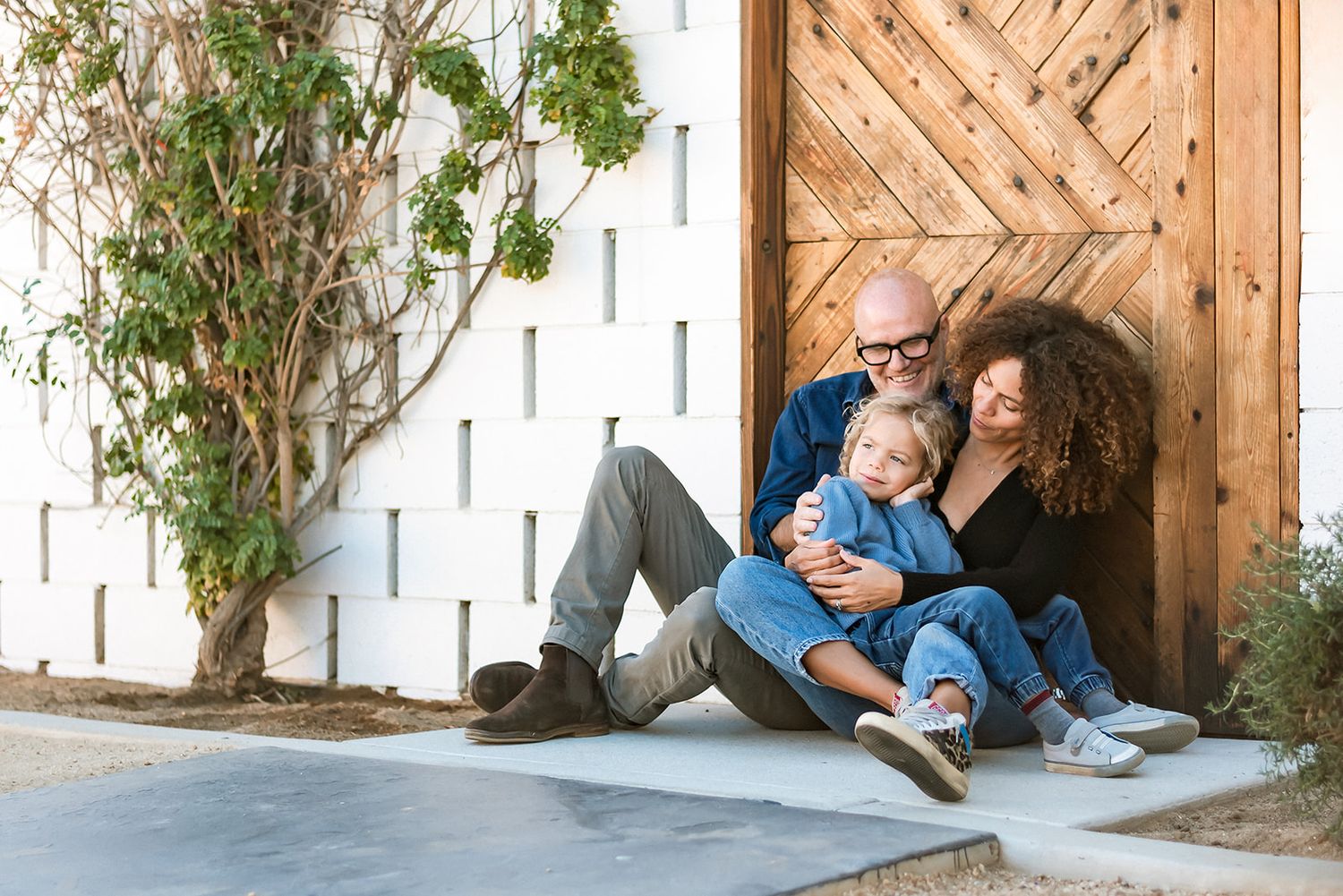 Happy family relaxing together in casual outfits while sitting by rustic wooden door with greenery climbing nearby.
