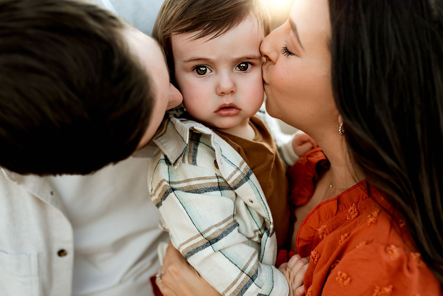 A small child in a plaid shirt receives loving kisses from both sides while being held during a photo session in Scottsdale