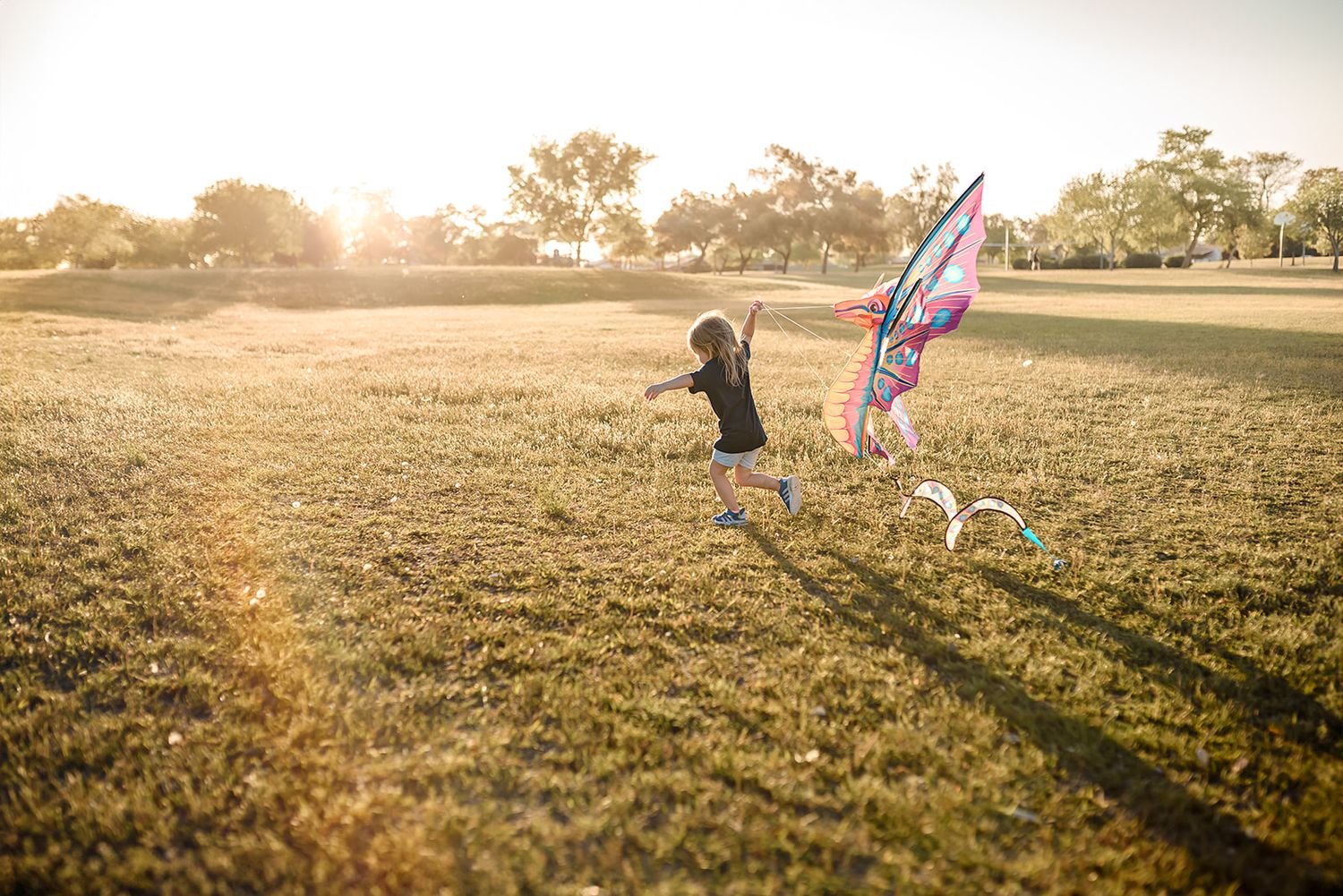 A little girl runs through a sunlit field with a kite trailing behind her at sunset during a fun family photo session in Scottsdale.