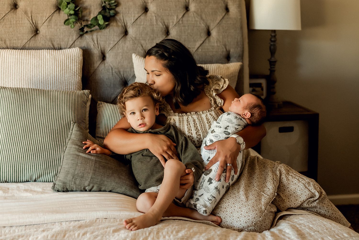Mom kisses toddler's head while holding her newborn baby on a bed during a lifestyle newborn photo session.