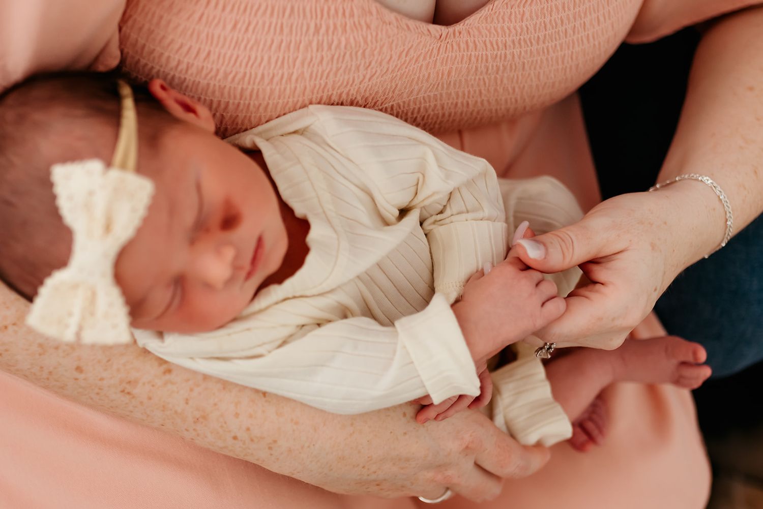 Close-up photo of a newborn baby in a cream-colored outfit with a bow, holding his mother's hand.