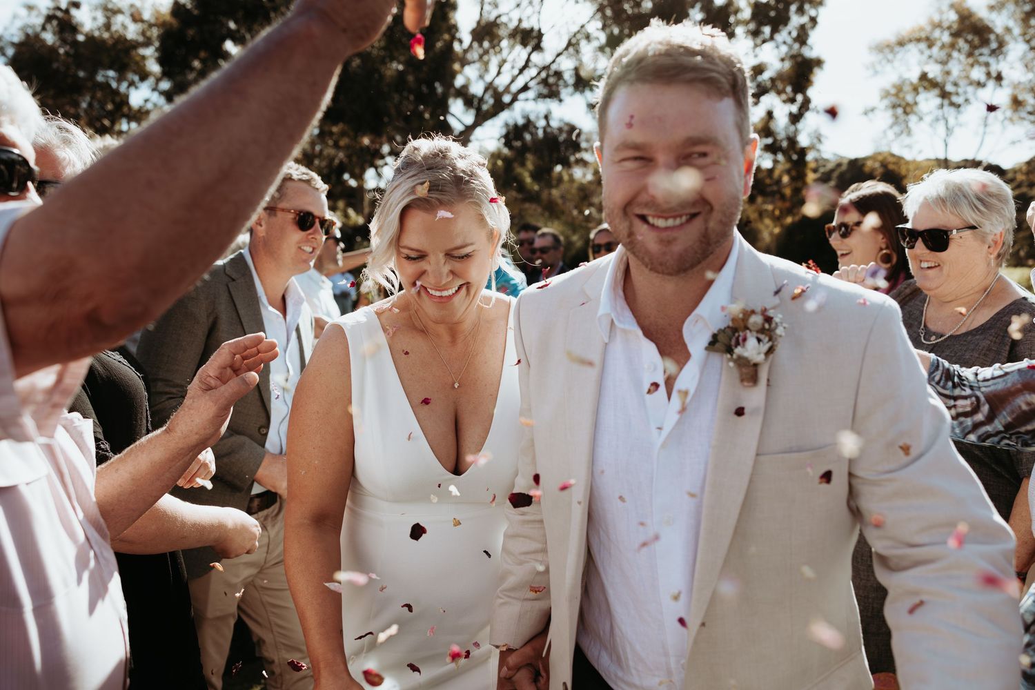 A group of wedding guests celebrate joyfully outdoors in white attire while red flower petals fall around them.