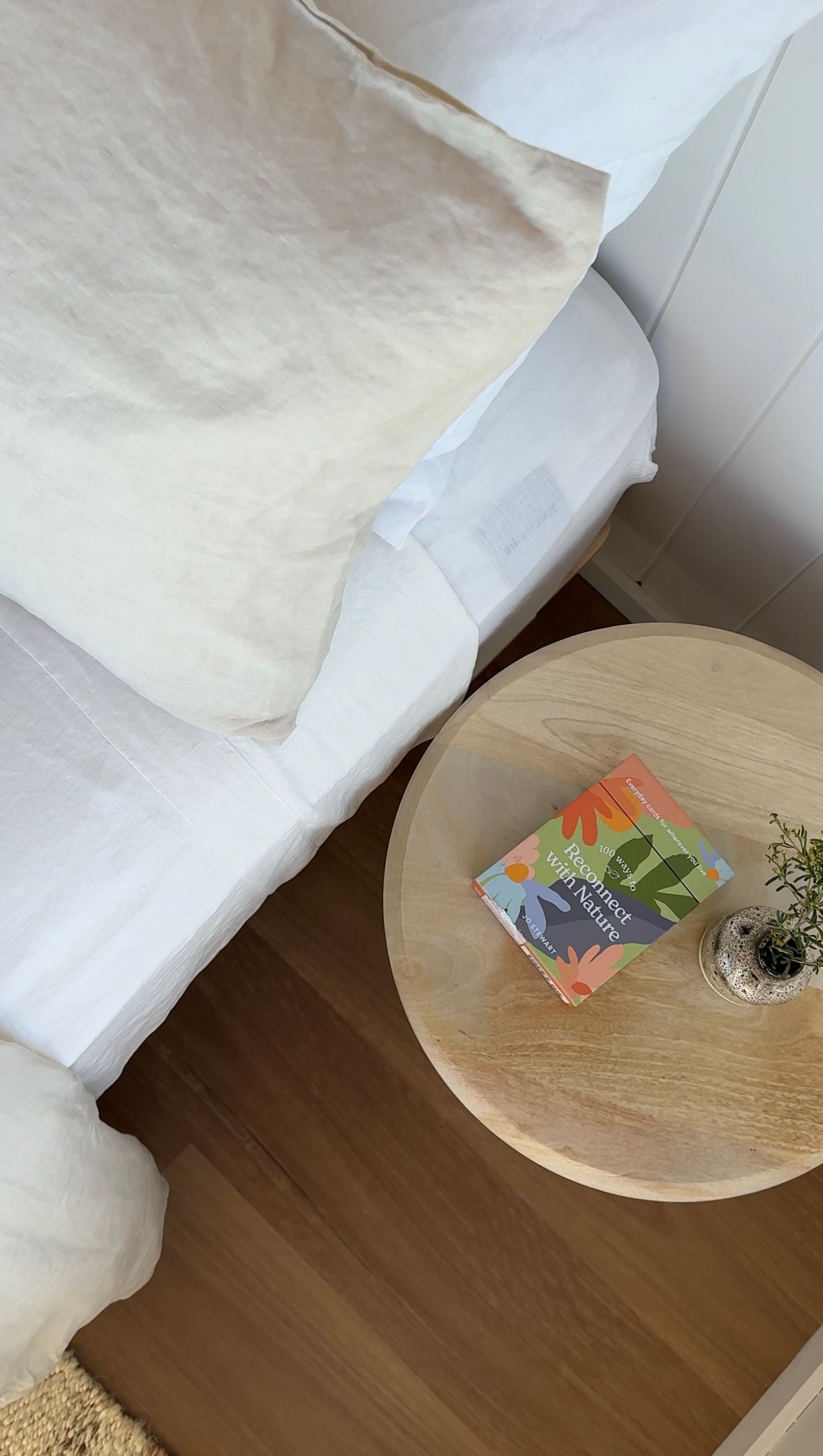 A round wooden side table sits next to a white bed or couch in a minimalist room with hardwood flooring.