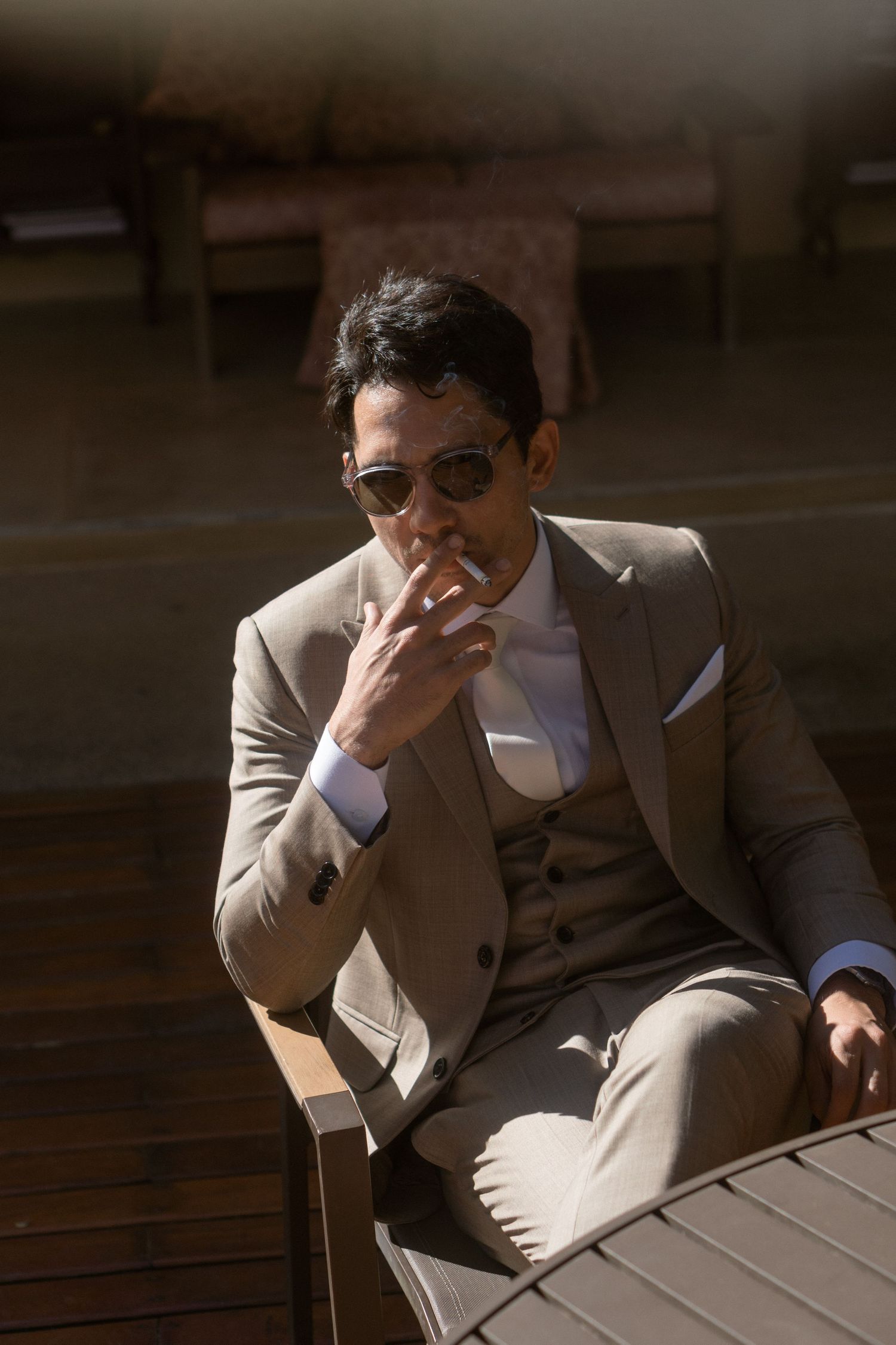 Um noivo estiloso vestindo terno bege e óculos de sol, fumando enquanto está sentado em mesa ao ar livre, em iluminação suave