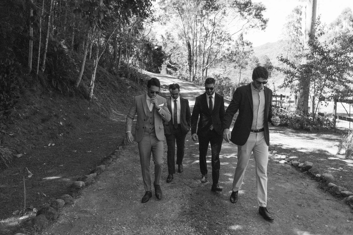 Foto em preto e branco do noivo e amigos caminhando por trilha em colina arborizada, a caminho da cerimônia para receber os convidados