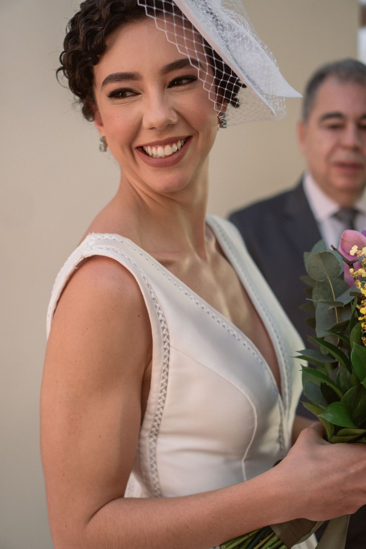 Noiva sorrindo com vestido de noiva branco sem mangas, véu vintage e buquê de flores naturais e galhos de eucalipto