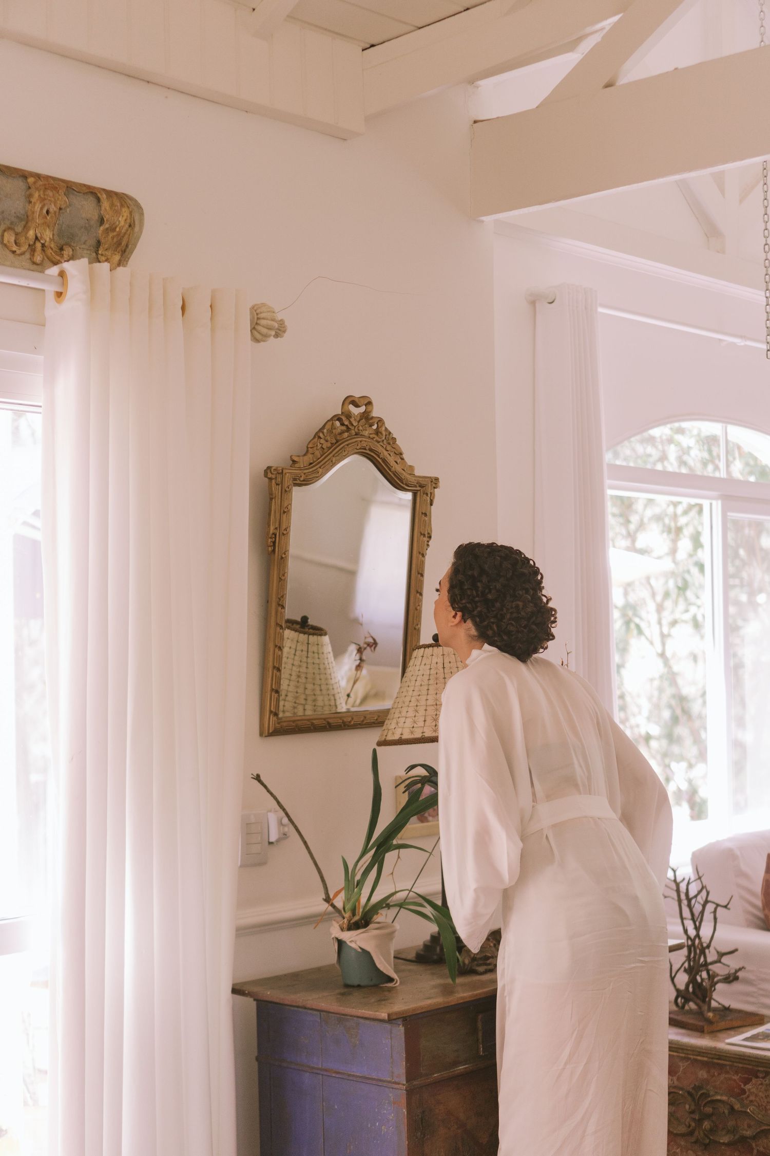 Noiva em roupão branco se preparando em um quarto iluminado e elegante, fotografado no estilo natural de Clara e Matheus