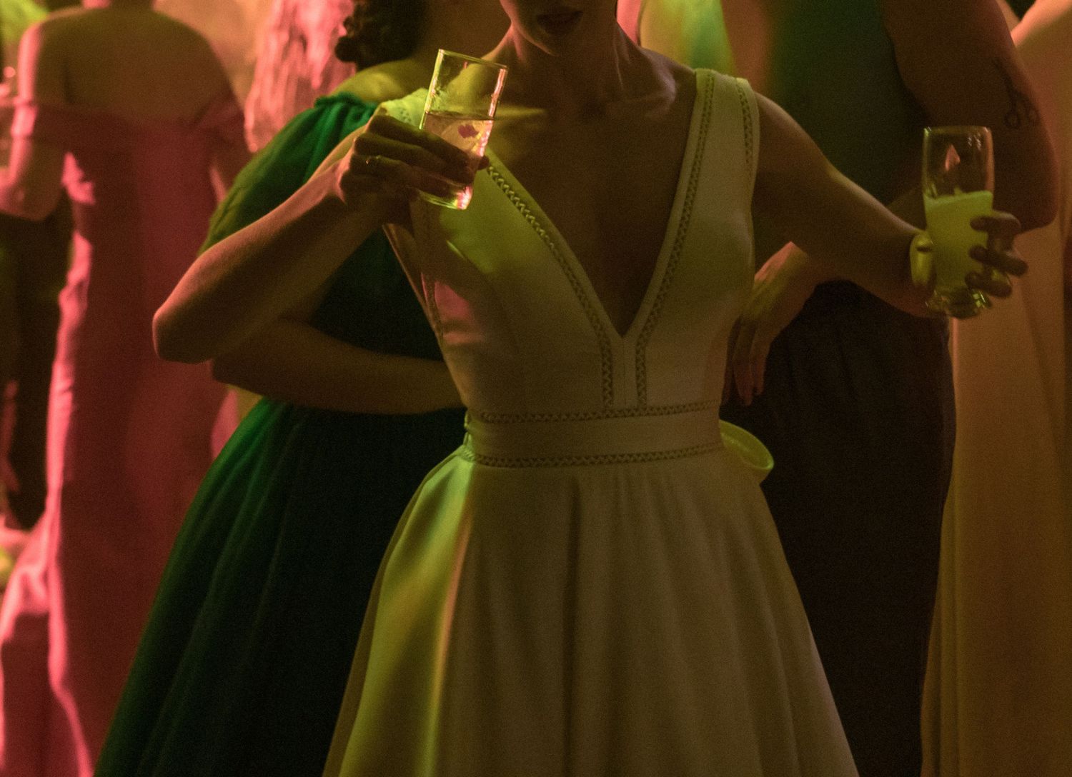 Noiva vestida de branco na pista de dança com drinks, sob luzes coloridas dramáticas, em uma fotografia artística e suave na festa casamento