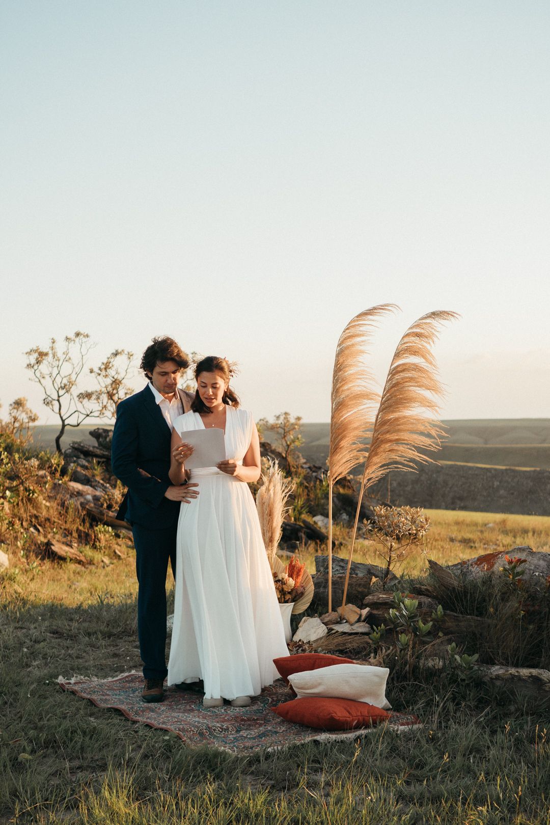 Um casal contempla o pôr do sol no topo de uma colina, com capim-dos-pampas e almofadas criando um cenário romântico ao ar livre.