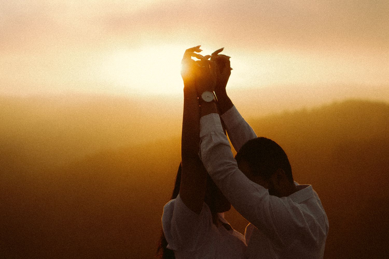 Silhueta de um casal de mãos dadas, erguendo as mãos contra um céu quente ao pôr do sol.