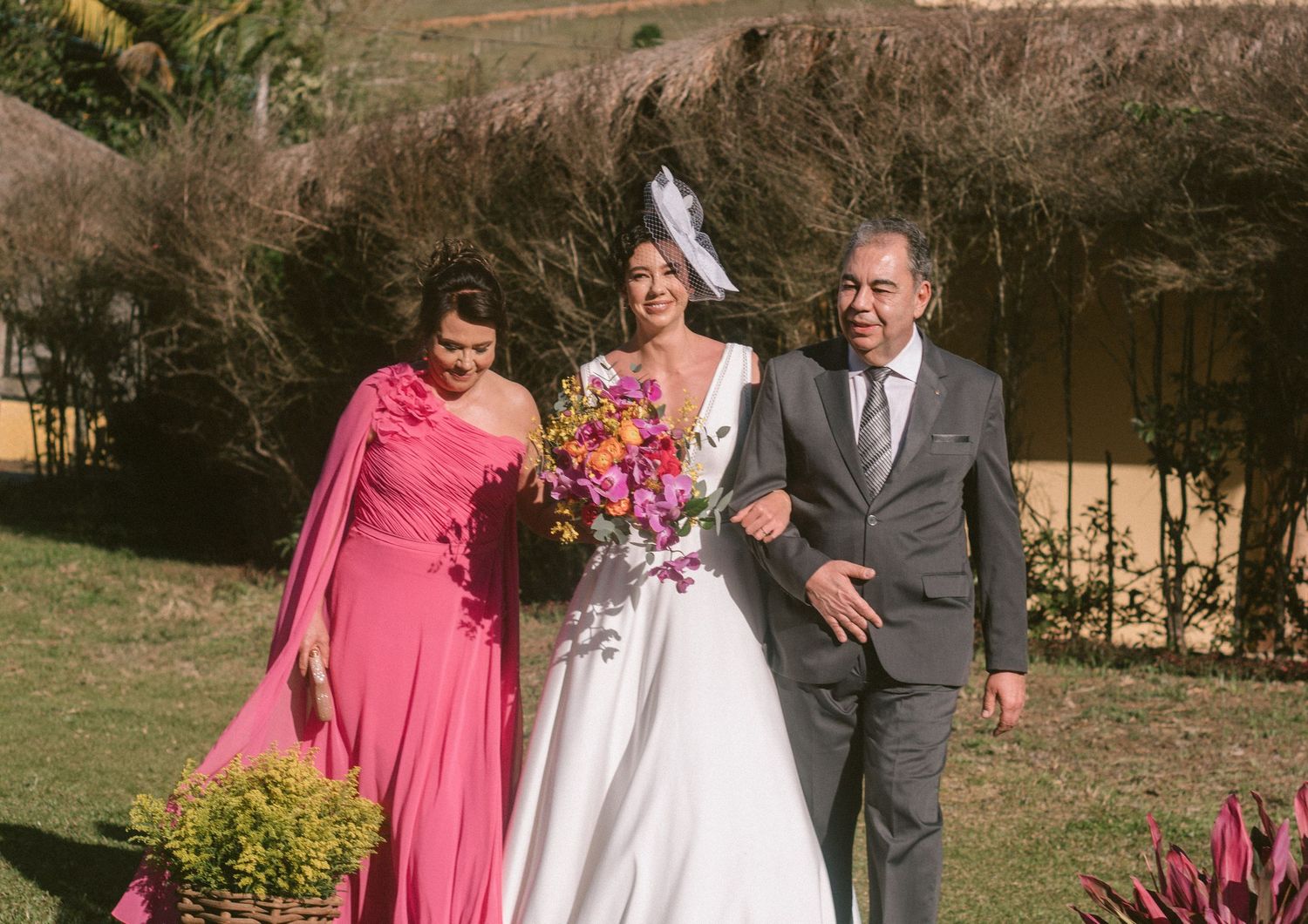 Noiva entra com os pais na cerimônia ao ar livre, com flores coloridas e vegetação ao fundo, em um casamento no jardim