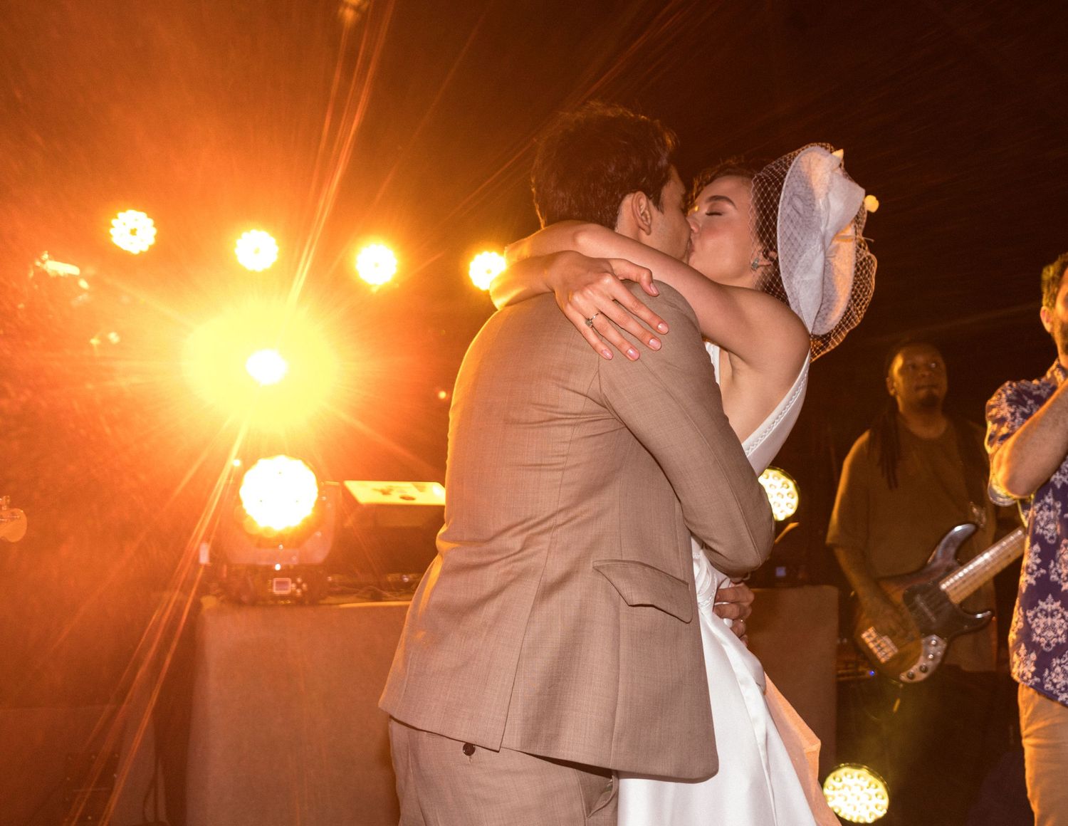 Casal se abraça na pista de dança da recepção de casamento, com luzes quentes e brilhantes ao fundo criando um clima acolhedor