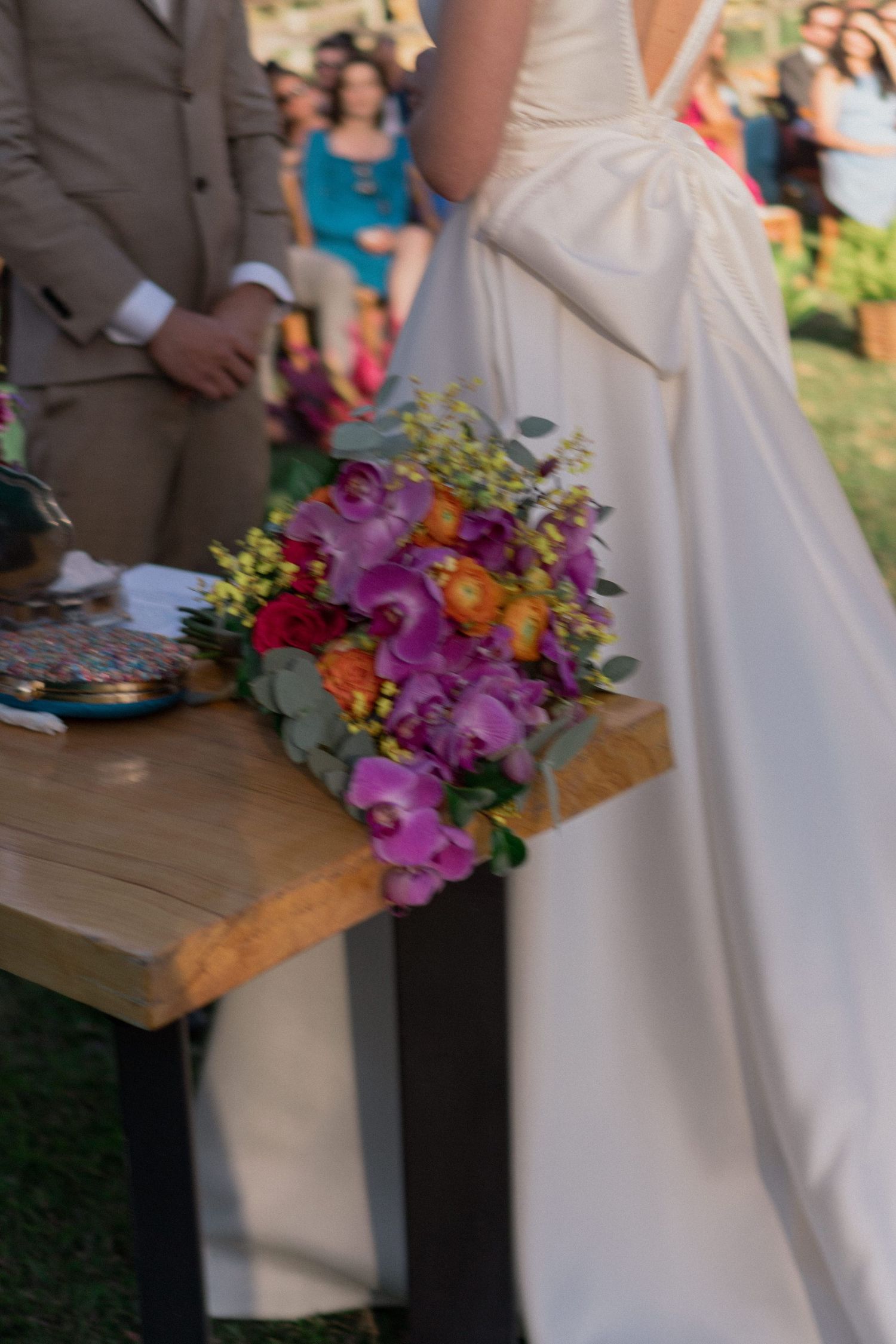 Detalhe do buquê da noiva, com orquídeas roxas e flores laranjas, complementando a beleza da cerimônia ao ar livre
