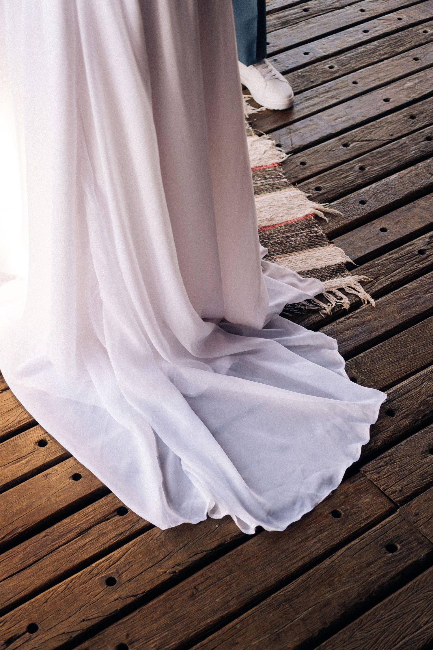 Cauda esvoaçante de vestido de noiva branco elegantemente drapeado sobre tábuas de madeira do deck.