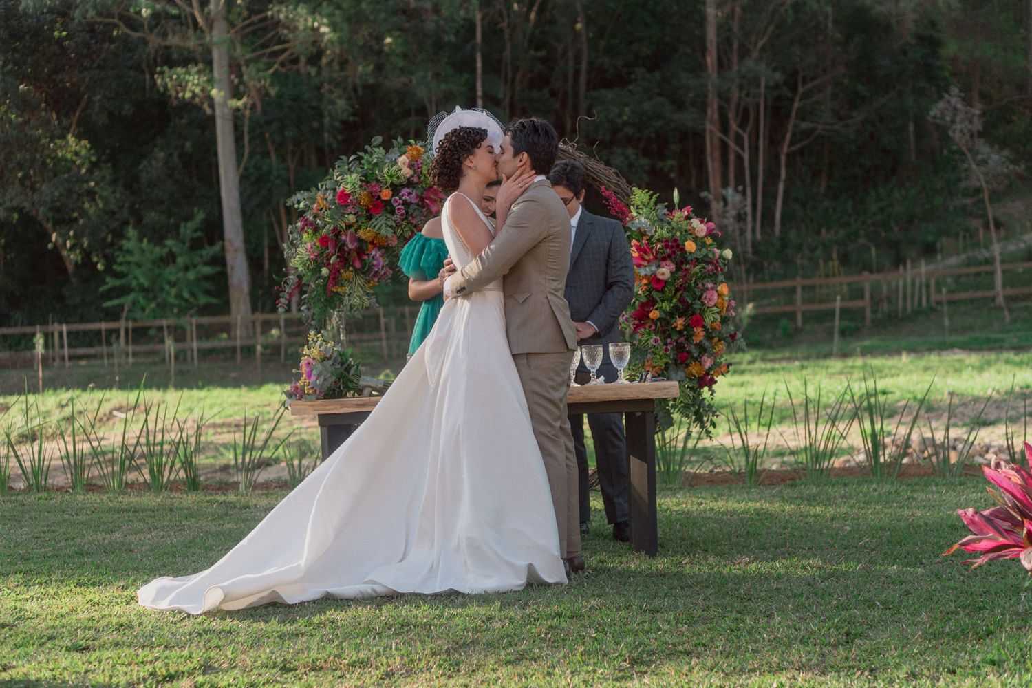 Casal se beija ao fim da cerimônia de casamento, sob um arco floral, em um ensolarado jardim ao ar livre Clara e Matheus®