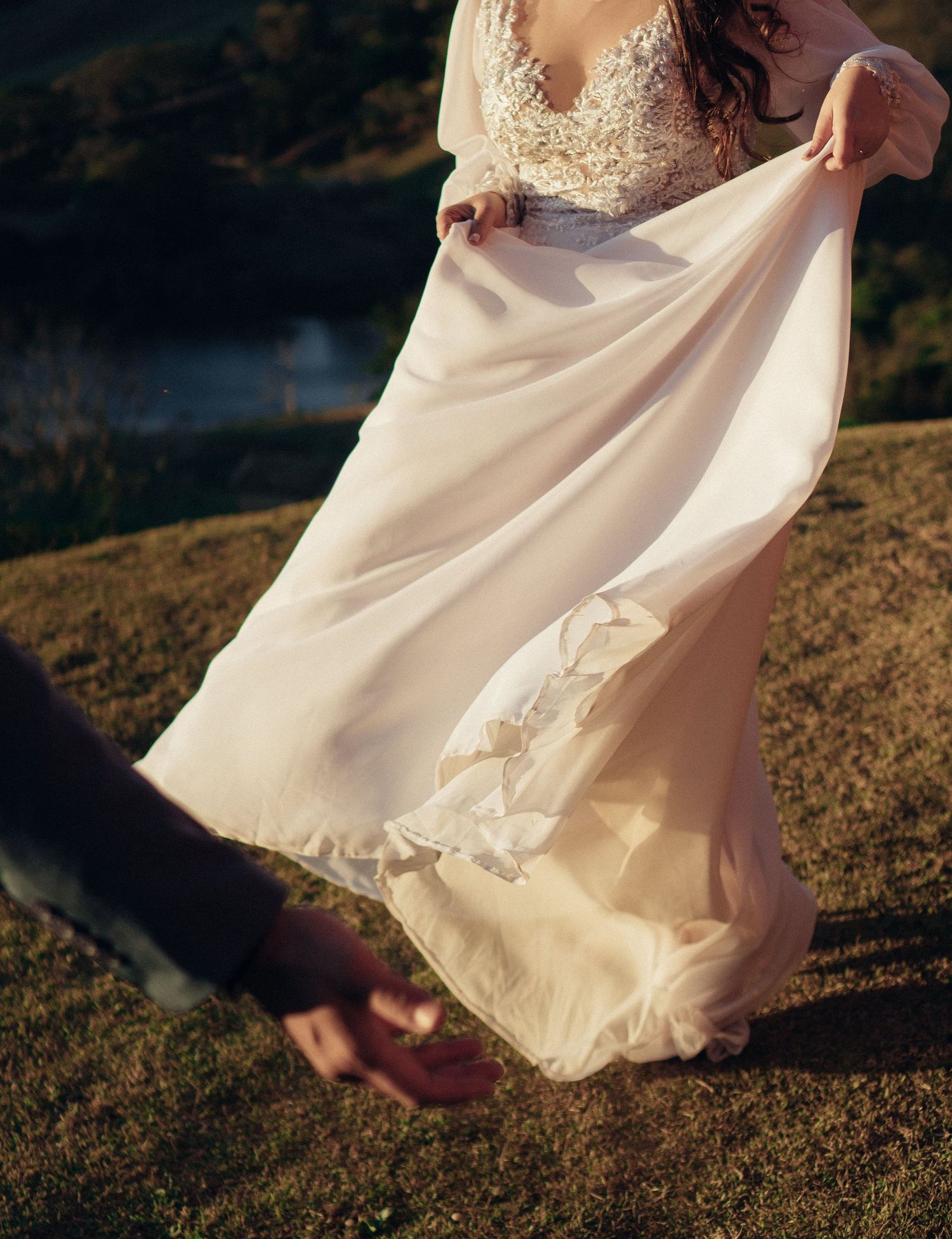Vestido de noiva branco esvoaçante com detalhes em renda captura a brisa com o cenário de montanhas ao pôr do sol.