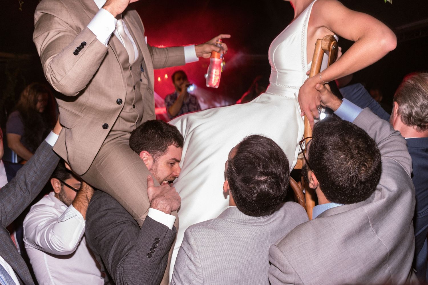 Noivos são levantados por amigos no meio da pista de dança, enquanto os convidados celebram animados sob luzes festivas em tons de vermelho