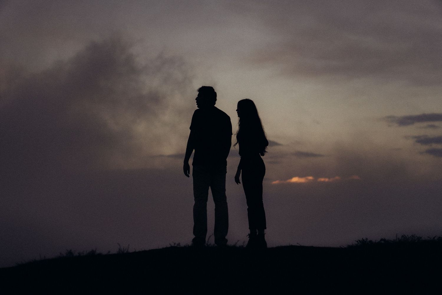 Silhuetas de duas pessoas em pé juntas contra um céu dramático de pôr do sol com nuvens escuras.