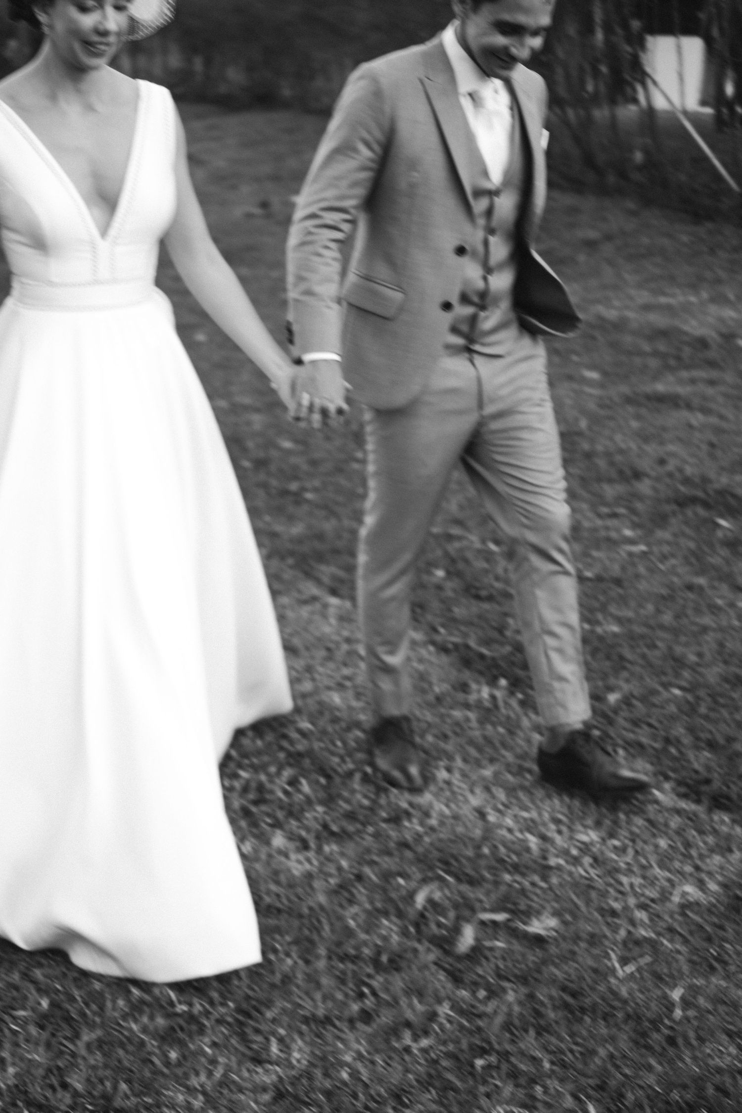 Casal de noivos elegante em uma foto de casamento preto e branco, caminhando de mãos dadas. A noiva usa um vestido branco fluído e o noivo u