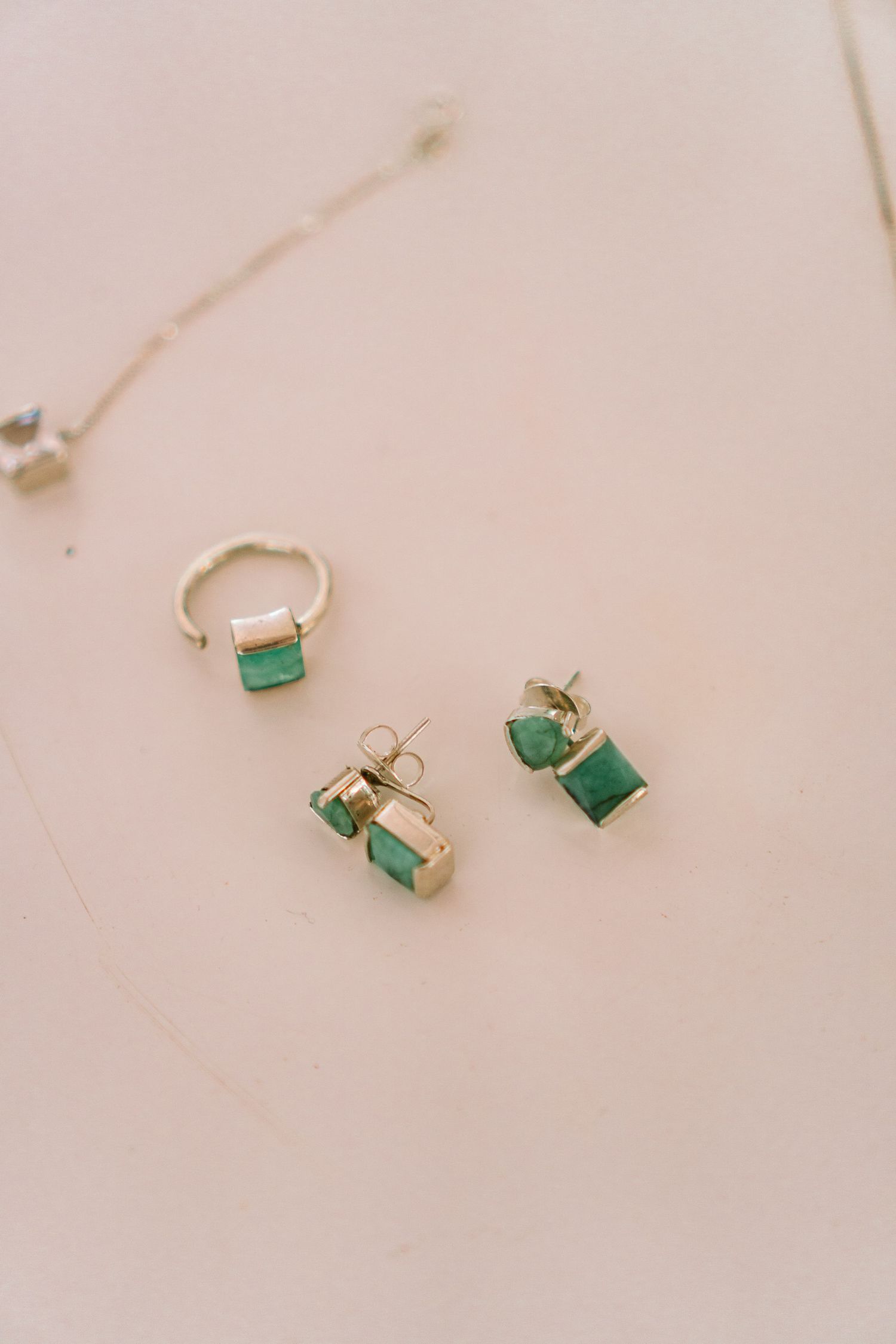 Conjunto elegante de joias douradas com pedras verdes esmeralda, incluindo anel e brincos de studs, sobre superfície rosa claro