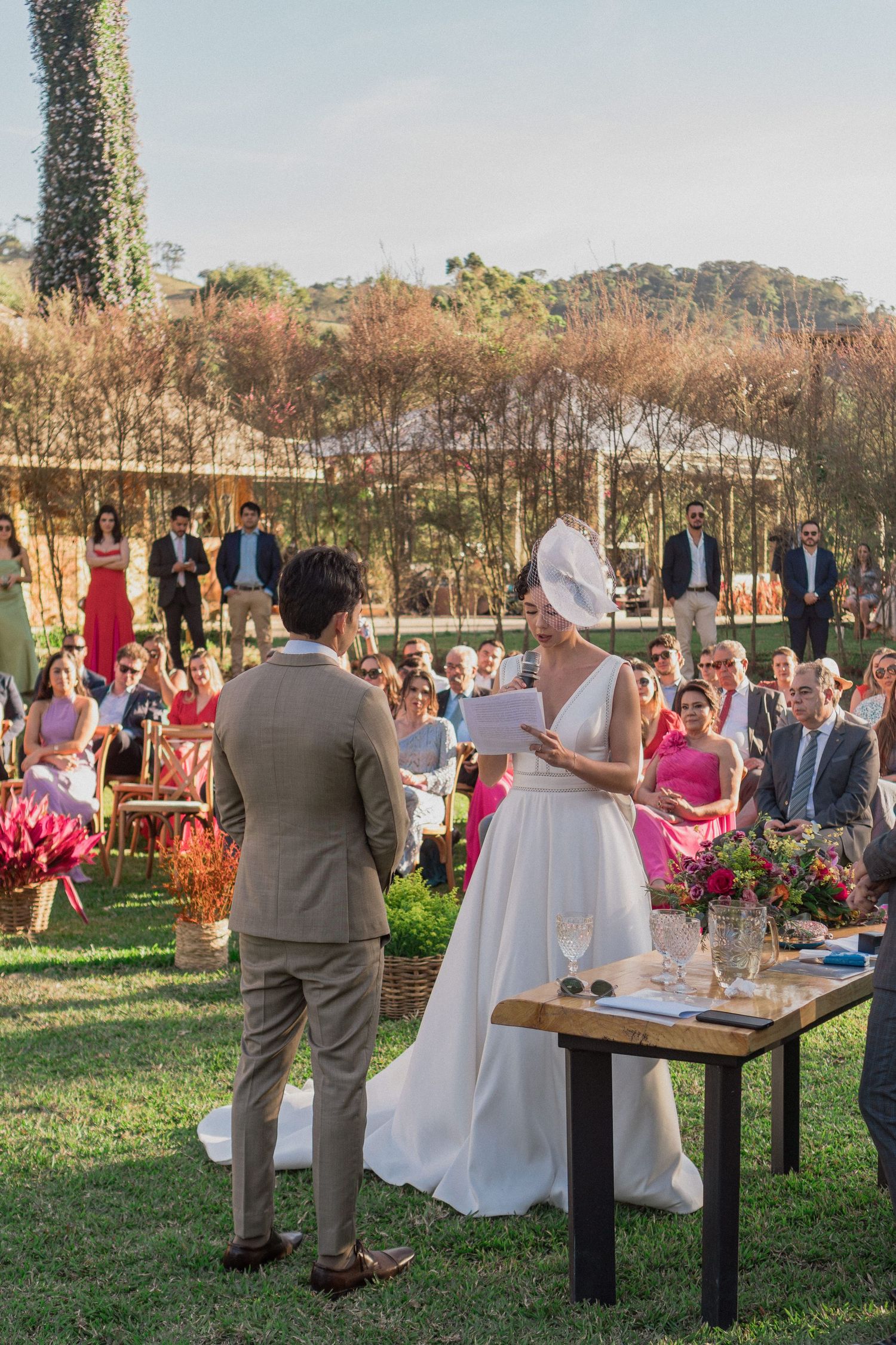 Cerimônia de casamento ao ar livre em um jardim, com os noivos lendo seus votos, rodeados por convidados estilosos com a luz do por do sol
