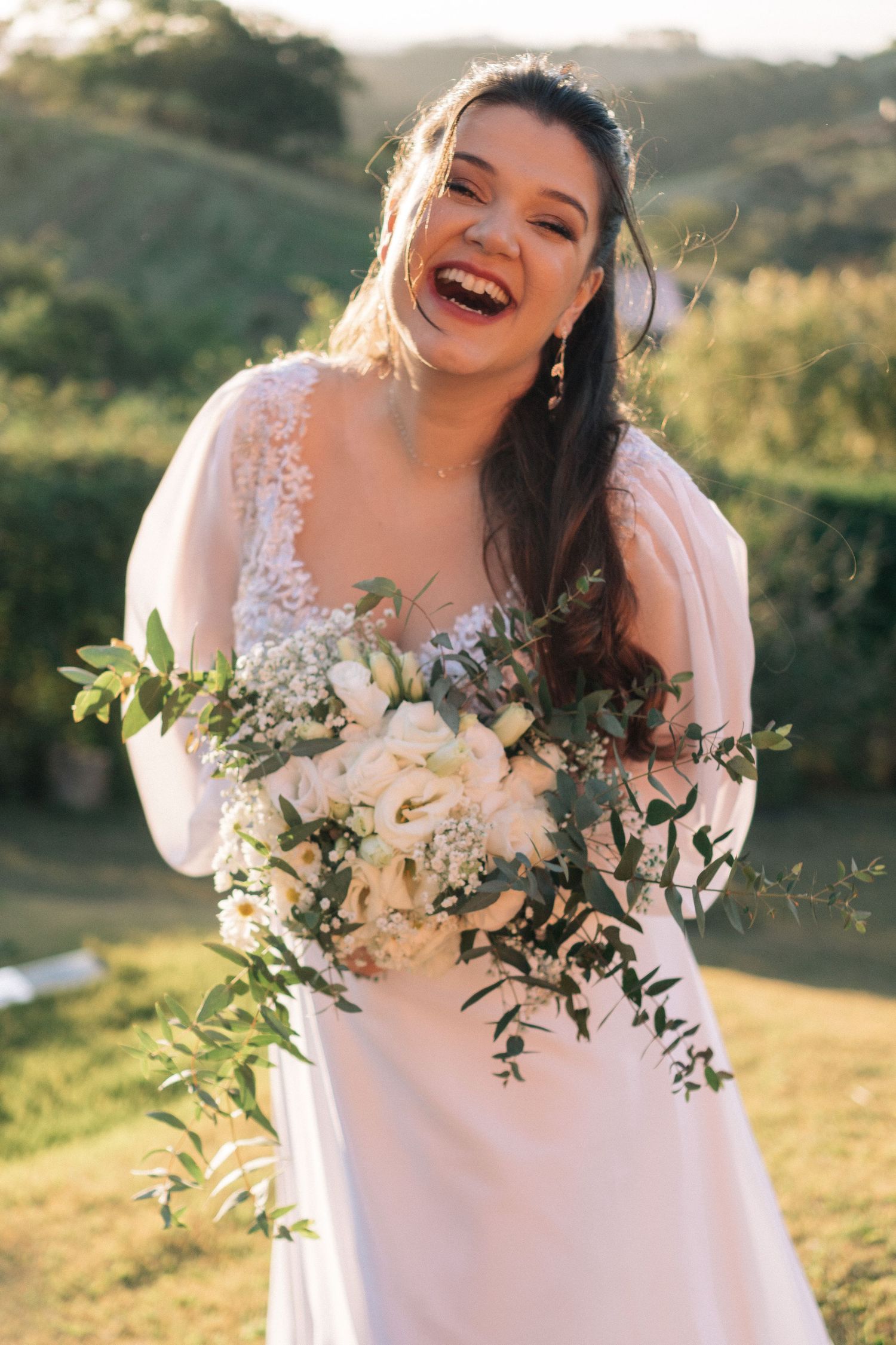 Noiva alegre em um vestido branco com detalhes em renda segura um buquê em cascata de flores brancas e folhagens em uma encosta ensolarada.