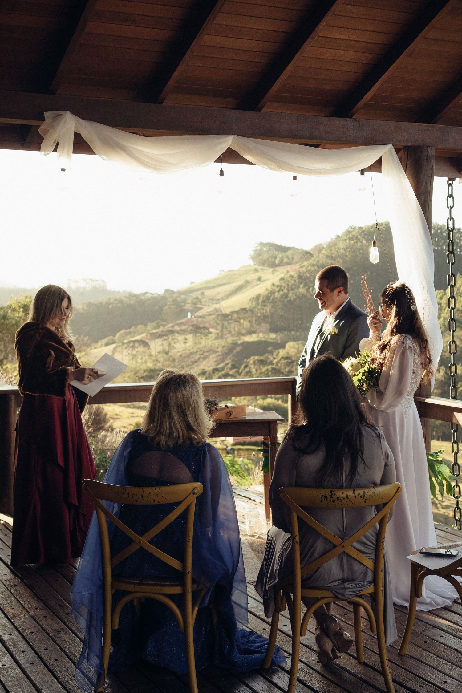 An intimate wedding ceremony takes place on a rustic deck overlooking scenic rolling hills at sunset.