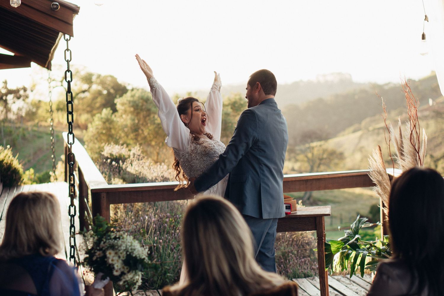 Celebração de casamento em deck ao ar livre com montanhas e convidados ao fundo.