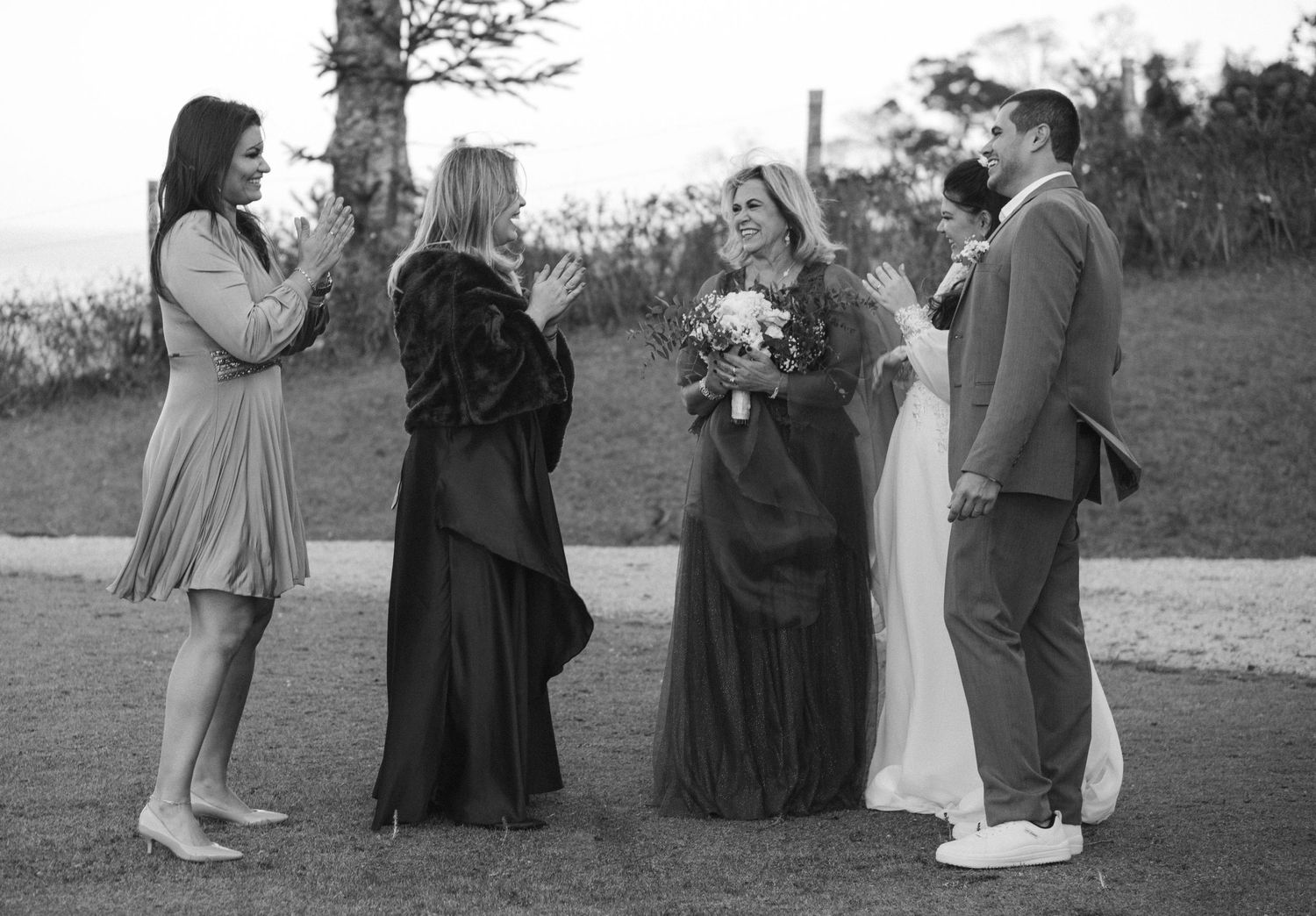 Uma foto em preto e branco captura os convidados do casamento compartilhando um momento emocionante ao ar livre.