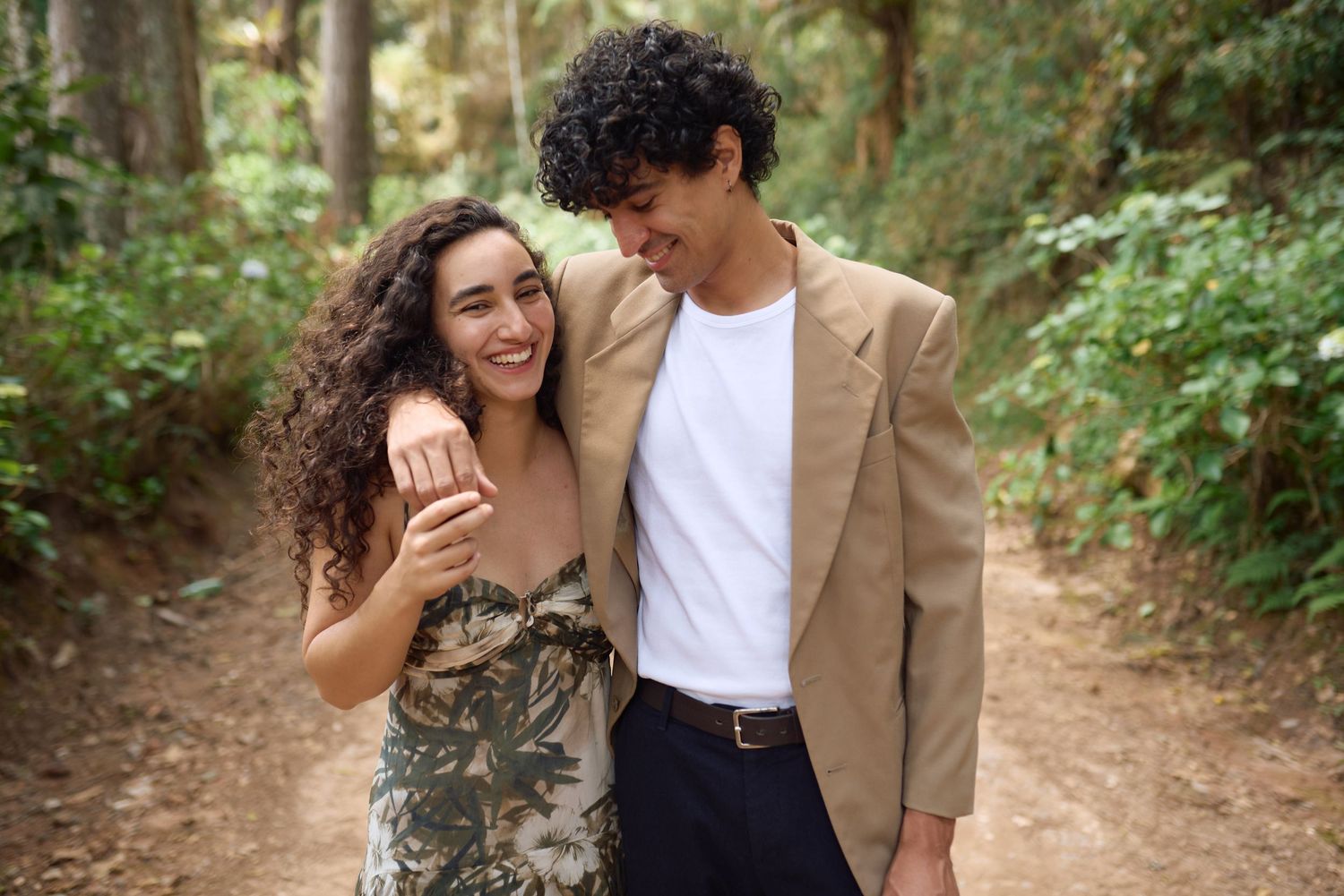 Um casal compartilha um momento terno enquanto caminha por uma trilha na floresta, cercado por vegetação exuberante.