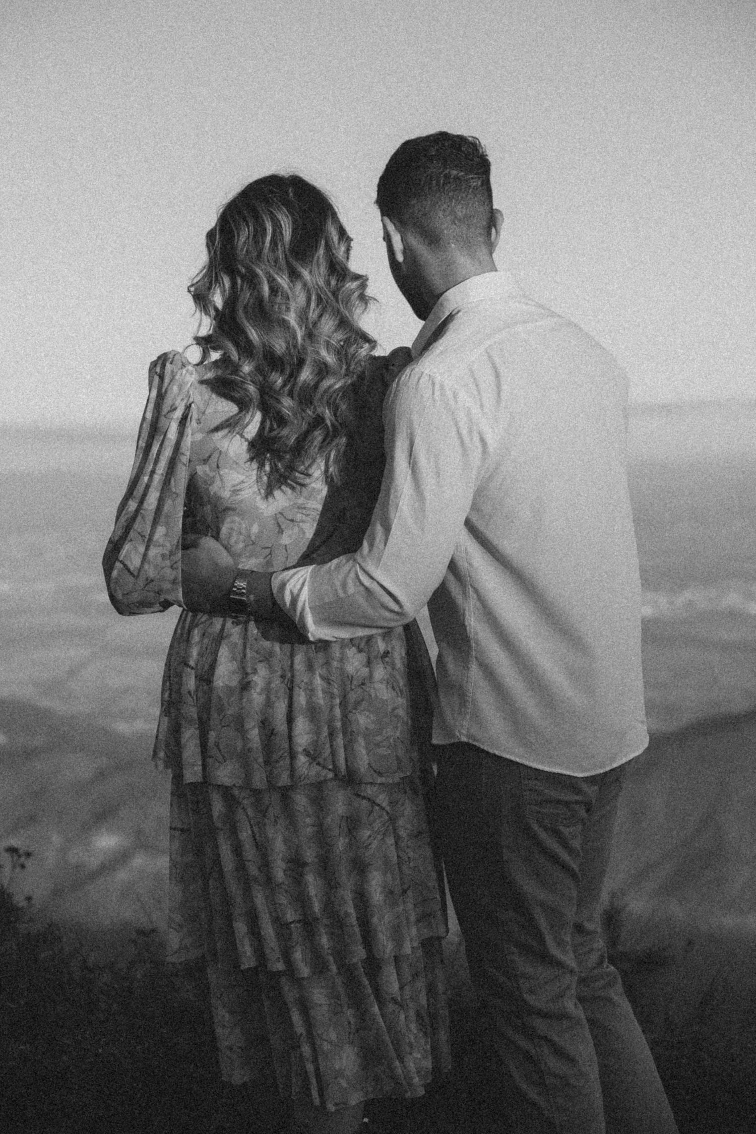 Um casal em silhueta posa junto contra uma paisagem montanhosa deslumbrante em fotografia a preto e branco.