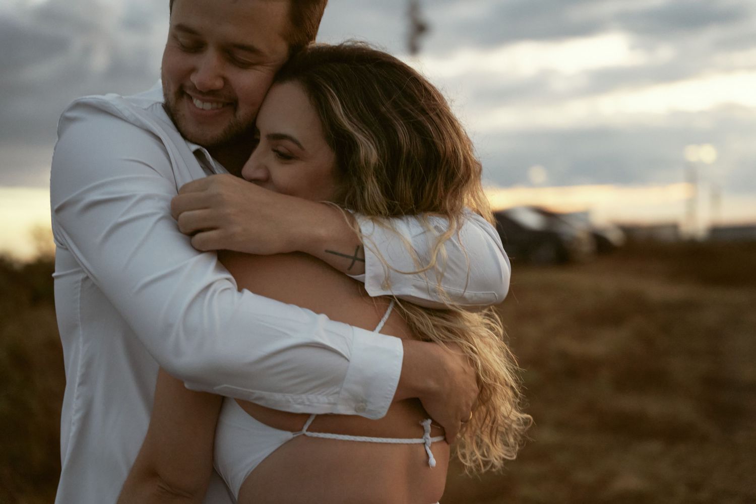 Um casal compartilha um abraço íntimo contra um céu nublado e melancólico ao pôr do sol.