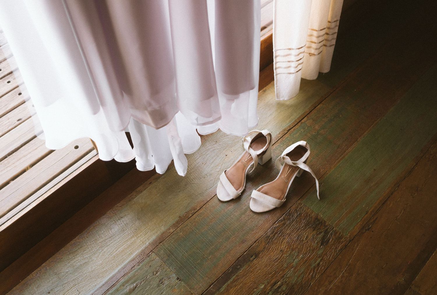 White wedding dress and elegant bridal shoes on wooden floor near window.