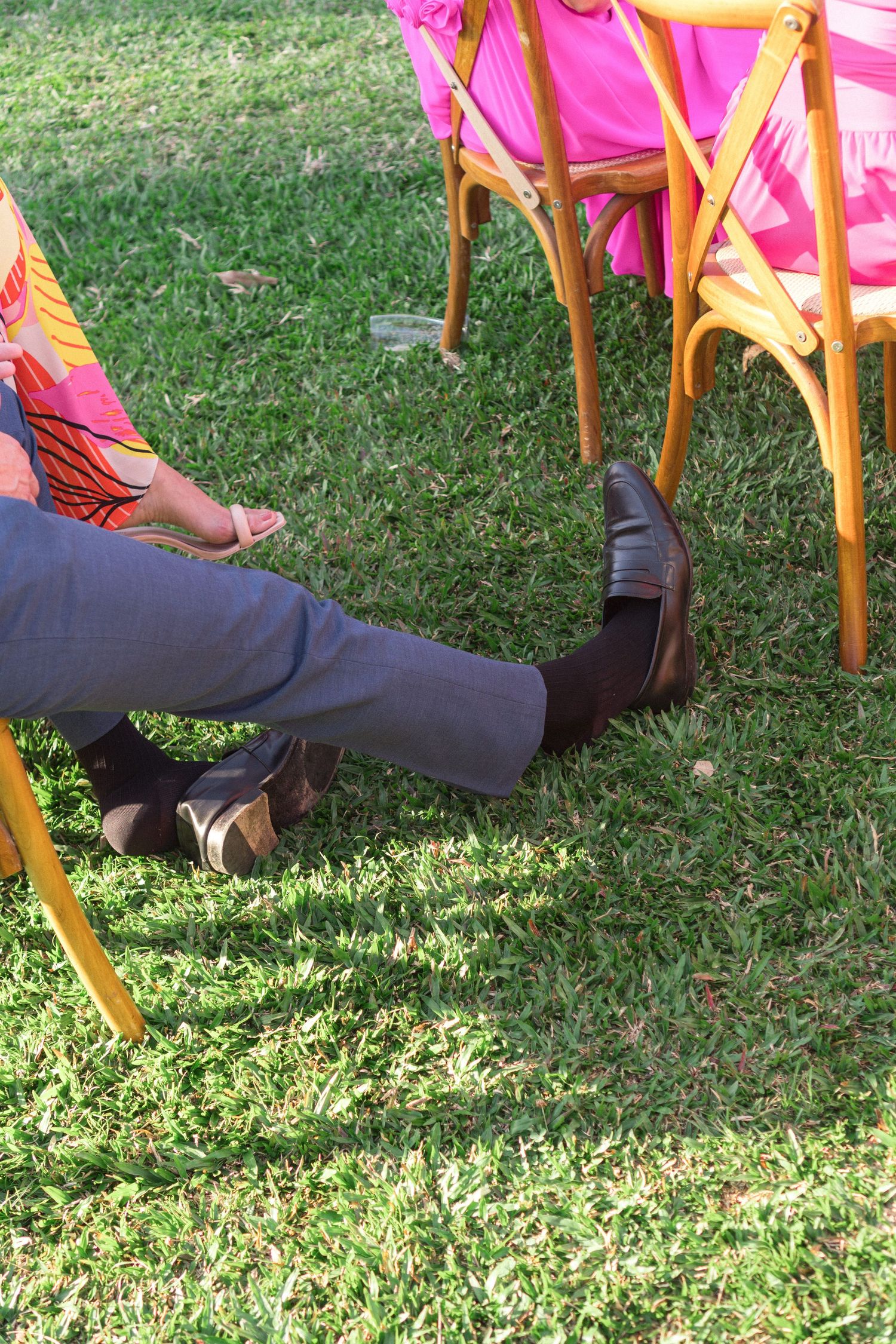 Detalhe de convidado relaxado durante a cerimônia de casamento, com calça cinza e sapatos marrons na grama ao ar livre