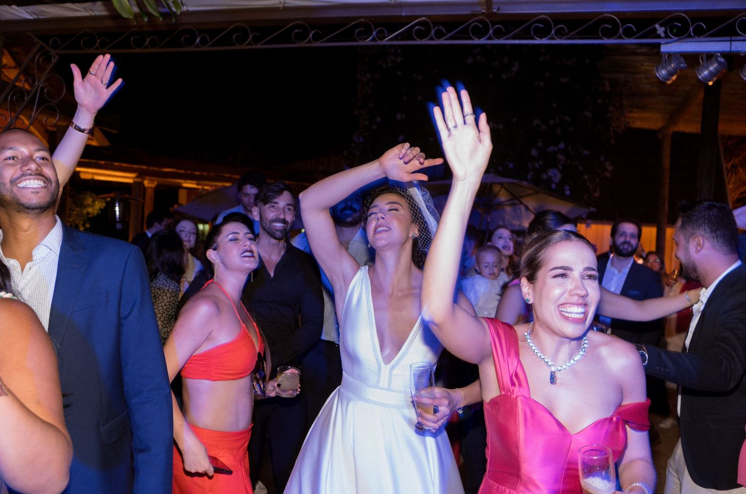 Convidados dançam e celebram com braços erguidos em uma pista de dança com iluminação suave durante a recepção noturna de casamento