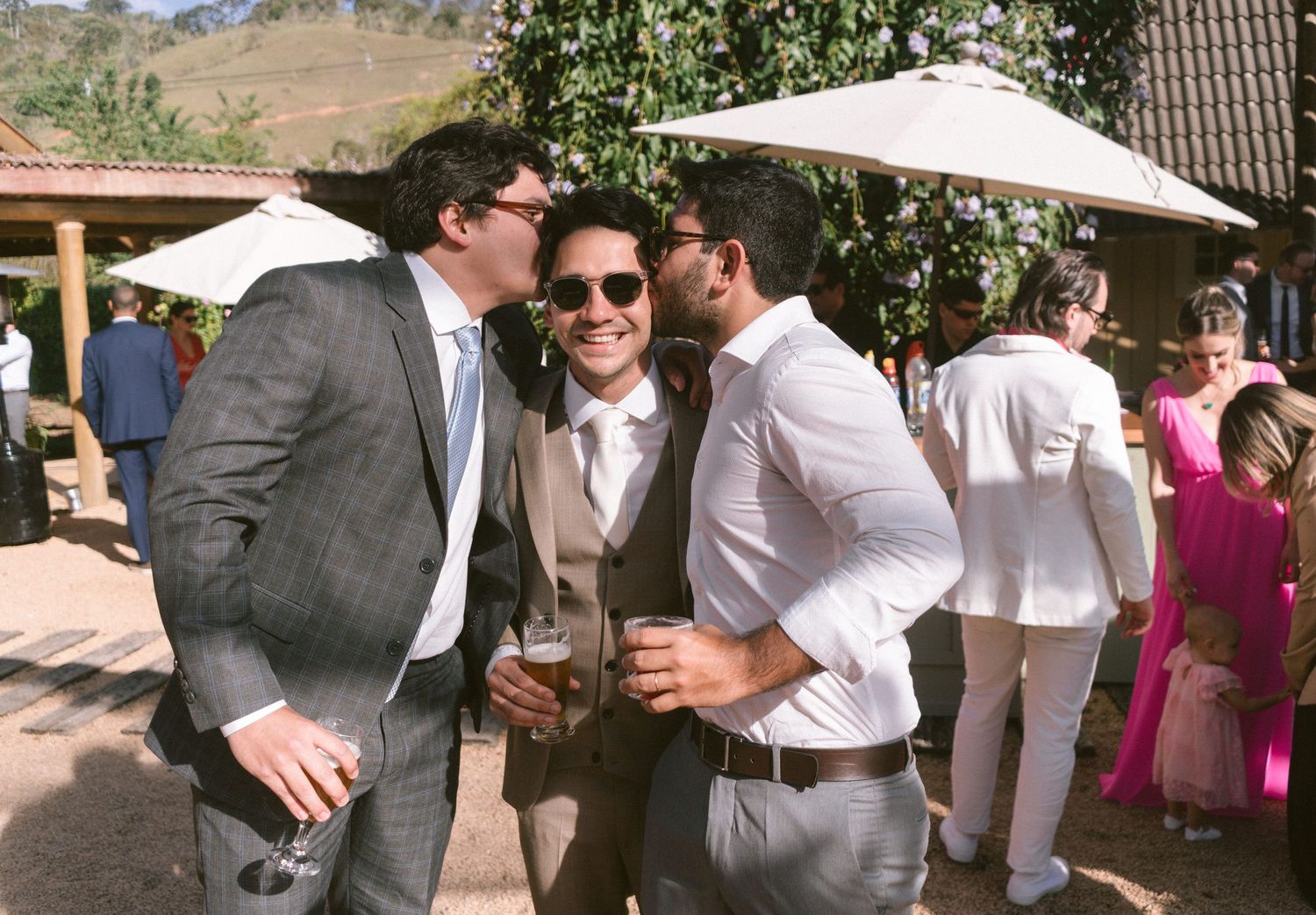 Três pessoas de terno e óculos de sol curtindo drinks em casamento ao ar livre, com guarda-sois brancos e convidados ao fundo