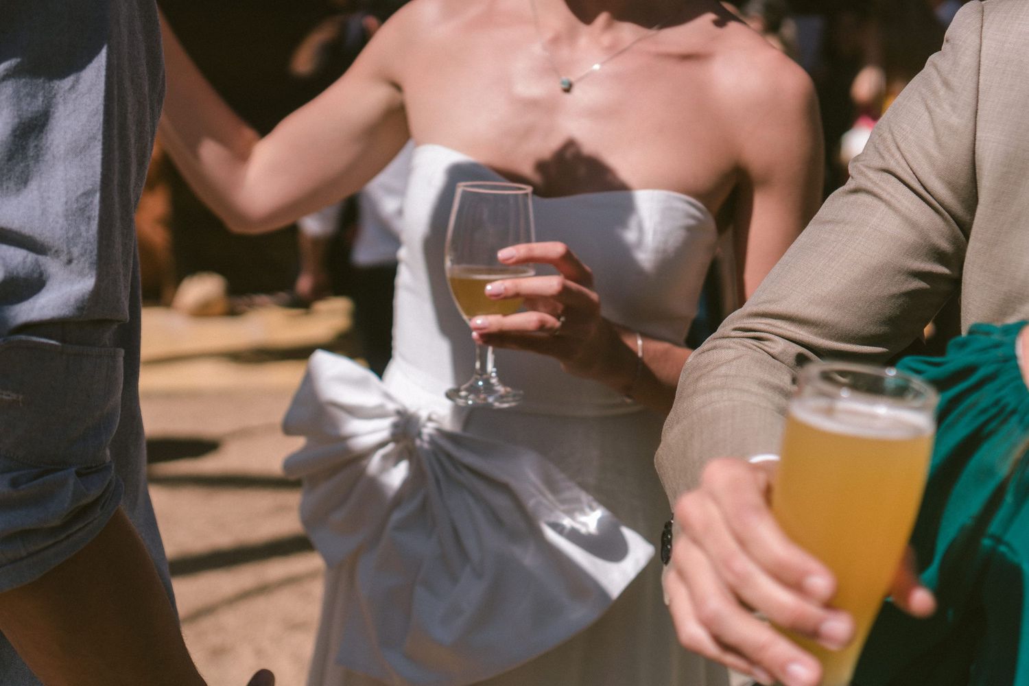 Noiva e noivo segurando drinks em um evento ao ar livre, com a noiva em vestido branco sob a luz do sol, fotografado em estilo documental de