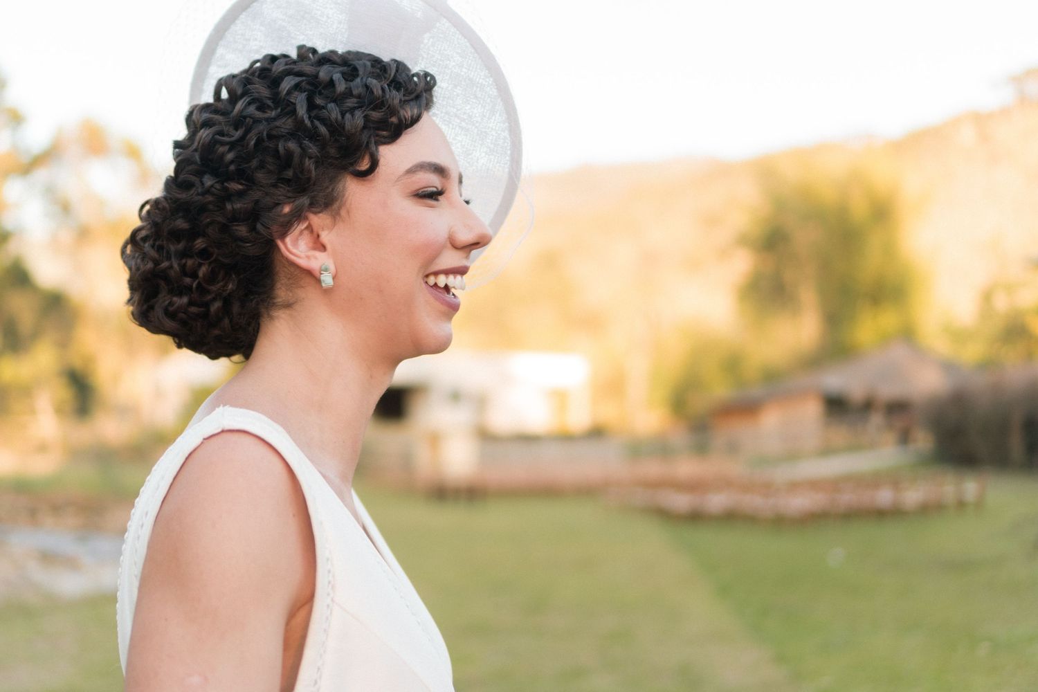Noiva de cabelos cacheados rindo alegremente em seu vestido branco, em um momento mágico ao pôr do sol