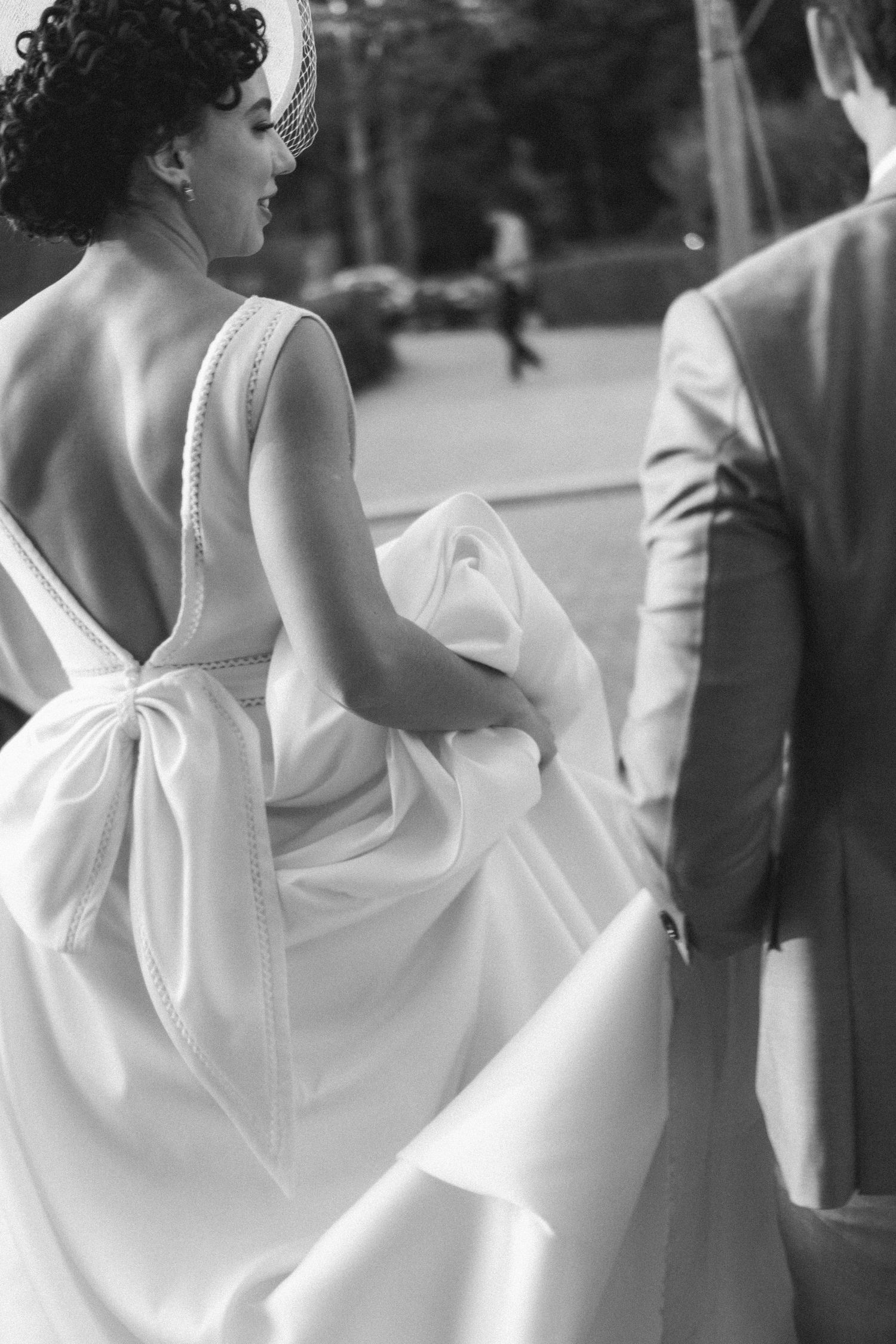 Foto em preto e branco da noiva com vestido branco elegante e laço nas costas, enquanto o noivo a ajuda a carregar o vestido