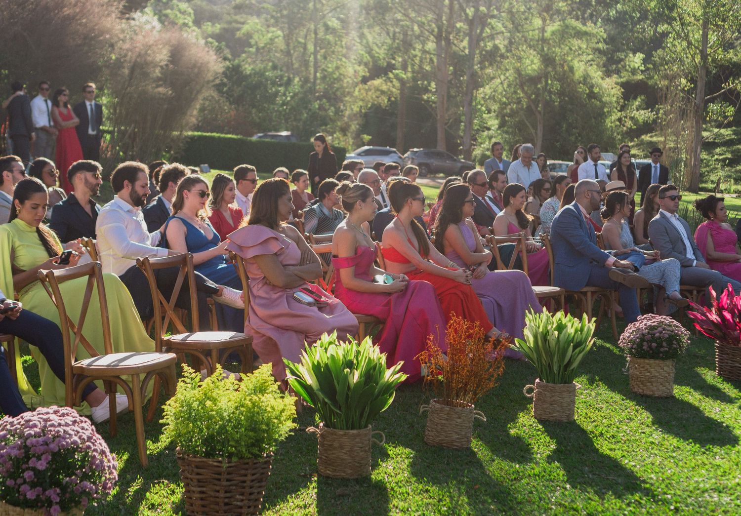 Detalhes encantadores: Convidados sentados em cadeiras de madeira, em uma cerimônia de casamento ao ar livre, com arranjos florais vibrantes