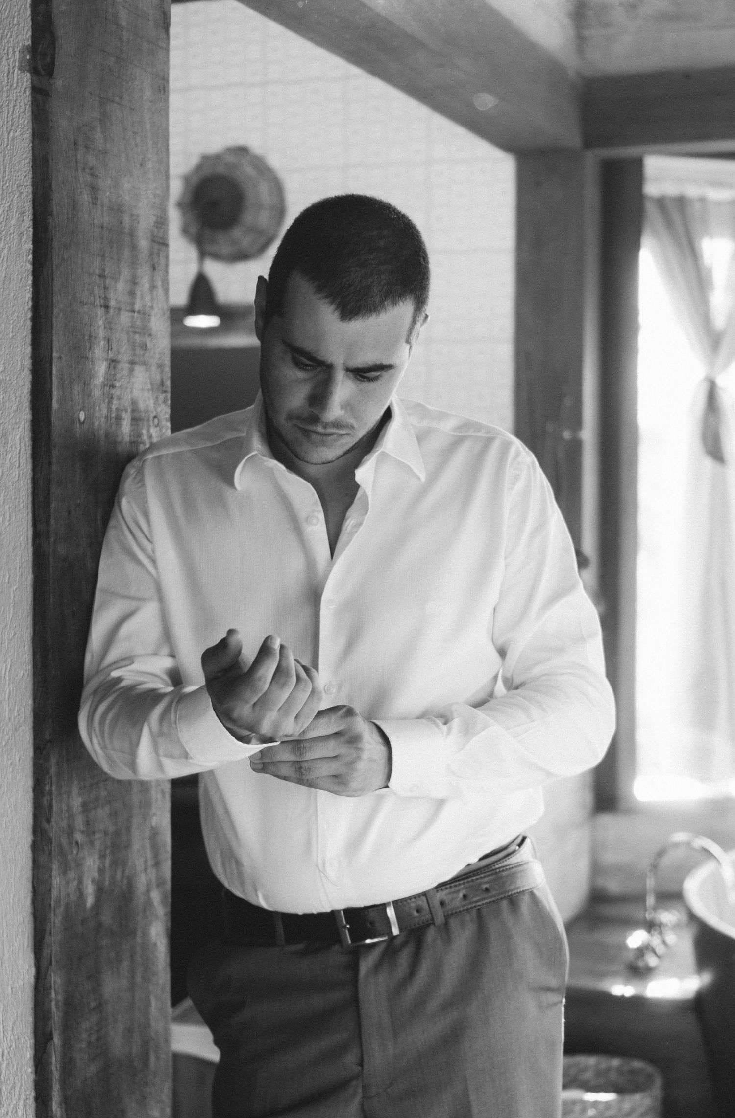 Fotografia em preto e branco de um noivo vestindo uma camisa social branca, ajustando o punho da camisa em um ambiente rústico.