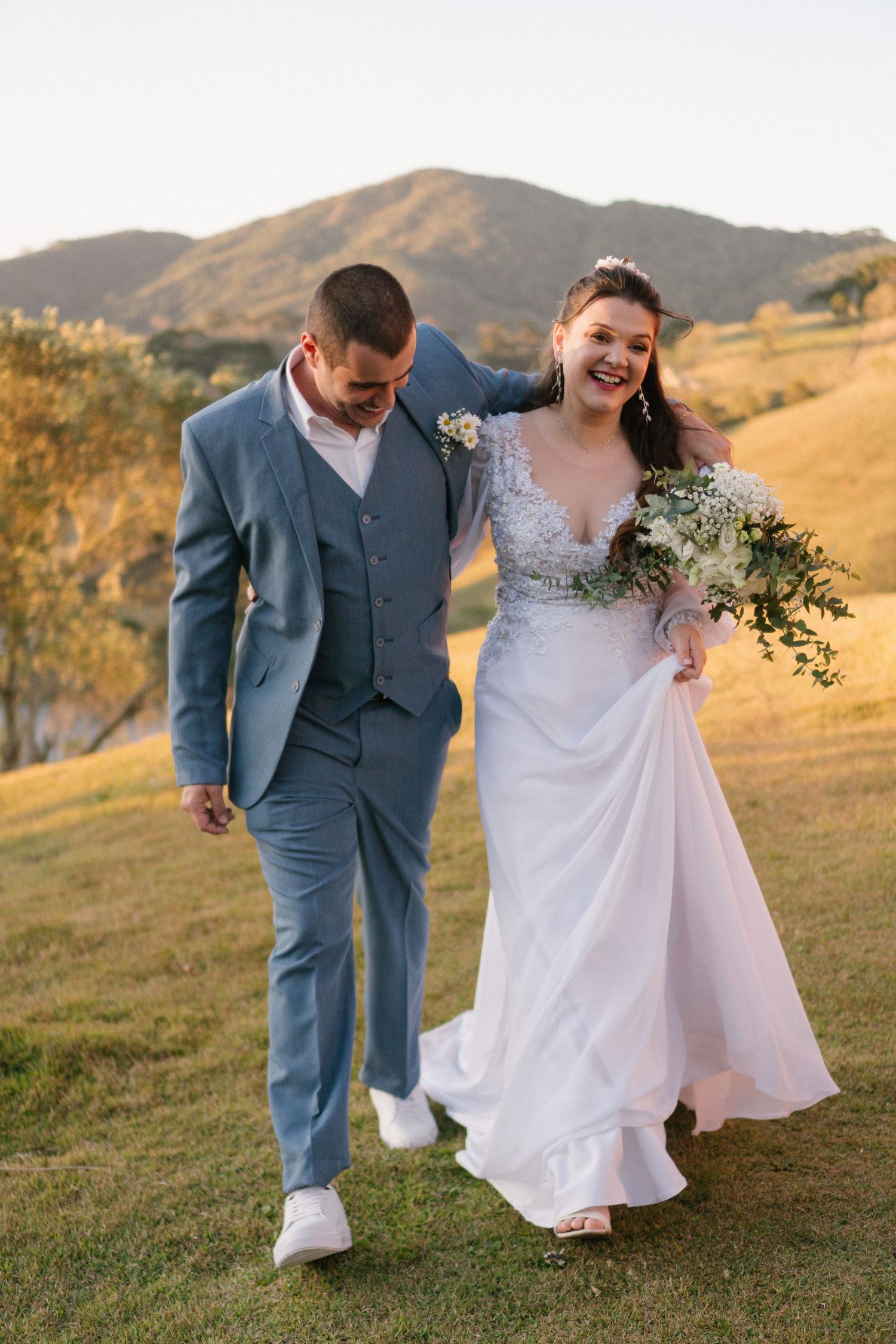 Recém-casados ​​felizes caminham juntos em uma encosta durante a hora dourada usando um terno azul e um vestido de noiva branco.