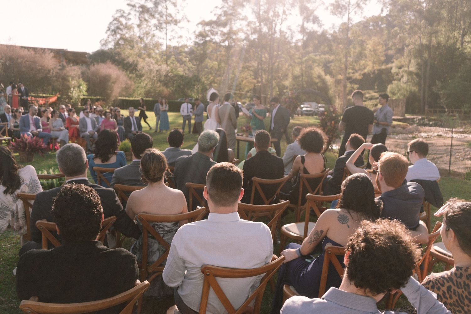 Cerimônia de casamento ao ar livre, com convidados sentados em cadeiras de madeira ao pôr do sol, luz dourada filtrando pelas árvores