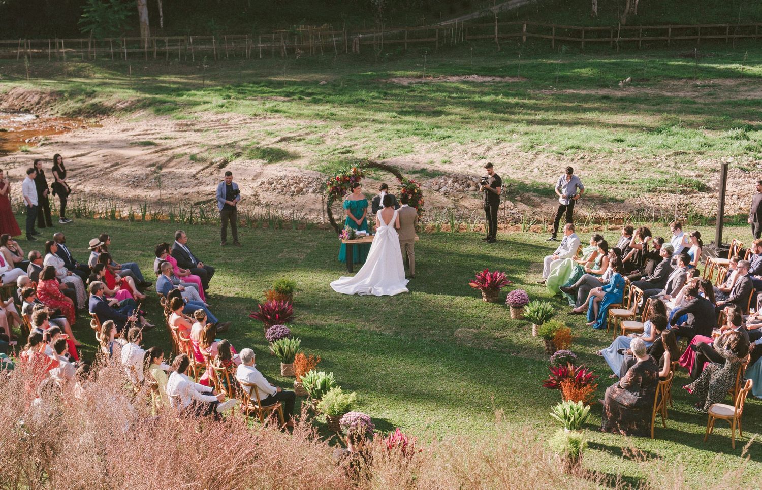 Cerimônia de casamento ao ar livre em campo gramado, com convidados sentados em cadeiras de madeira e arranjos florais coloridos ao redor fo